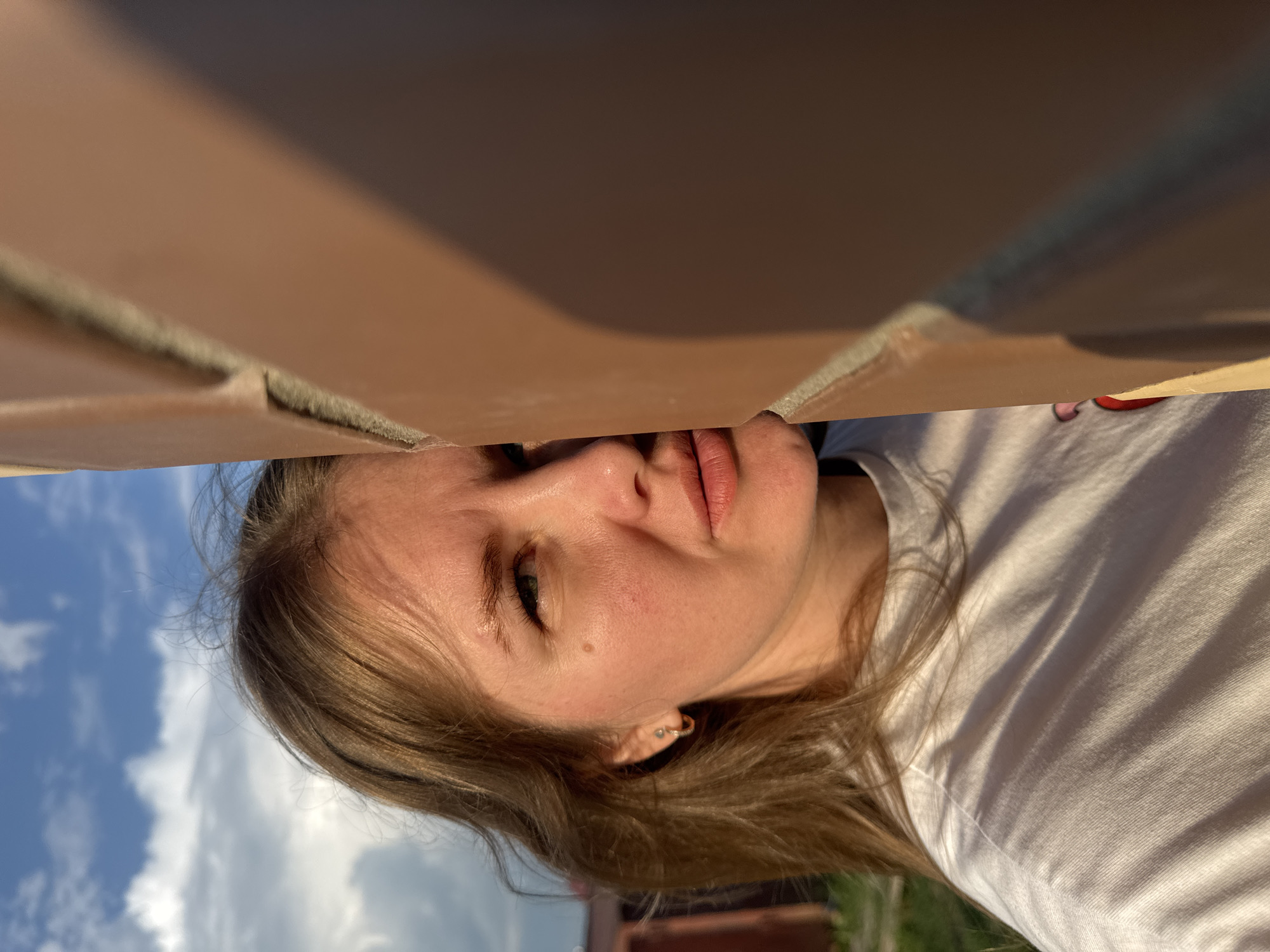A woman is positioned with a brick structure above her face. The background shows a partly cloudy sky and glimpses of greenery. The location could be Ферма, Russia, based on the prompt. The woman is wearing a white shirt. The bricks appear to be part of a wall or building element.