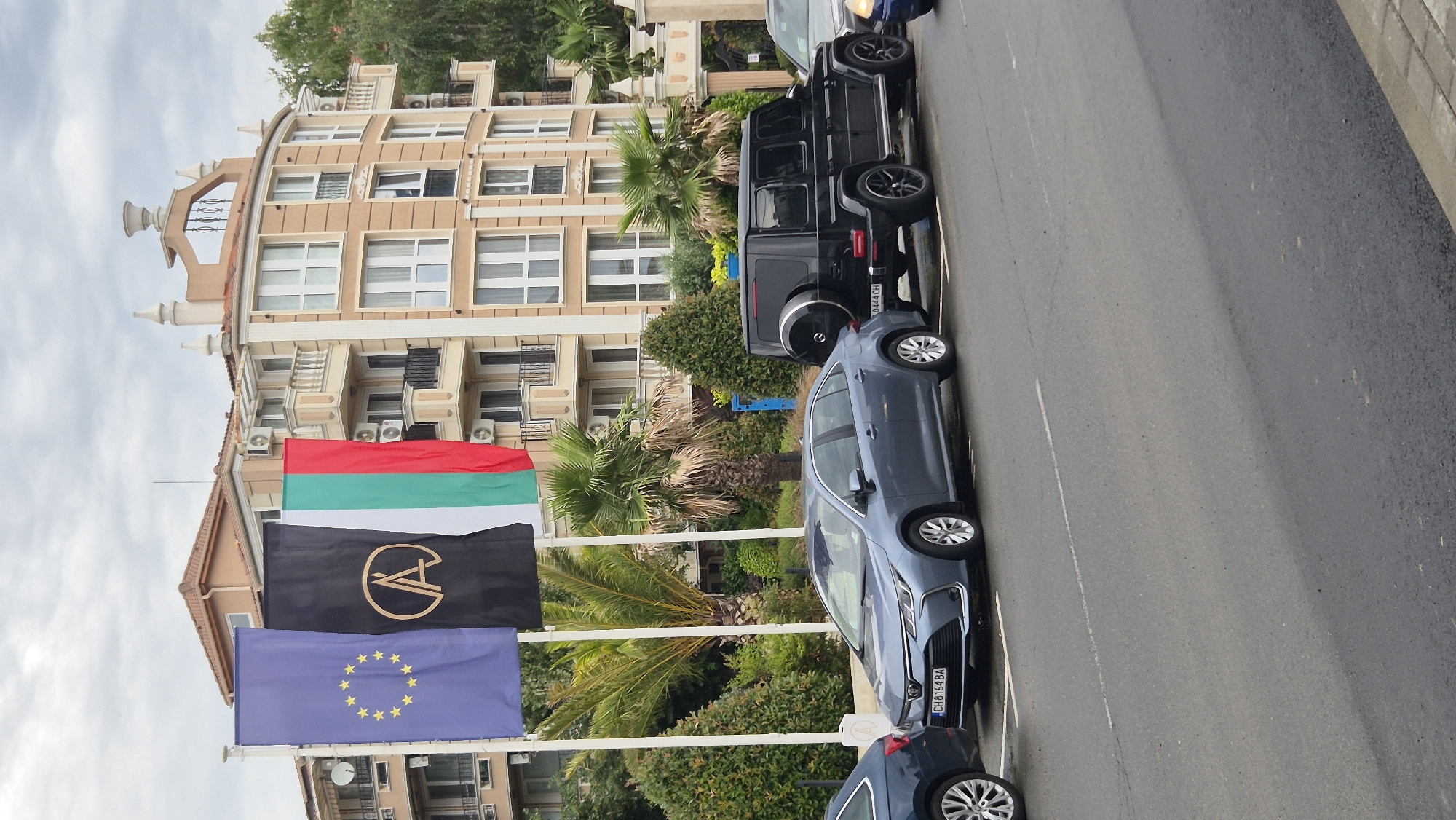 The image depicts a street scene in Ravda, Bulgaria, with a focus on a building and parked vehicles. Three flags are prominently displayed in front of a multi-story building. The leftmost flag is the flag of the European Union. Next to it is a black flag with a stylized golden symbol, possibly a logo. The rightmost flag is the Bulgarian national flag, featuring vertical stripes of white, green, and red.
The building is light beige with multiple balconies, white railings, and numerous windows. It appears to be a hotel or apartment complex. Tropical plants, including palm trees, are visible in front of and around the building, suggesting a coastal or warm climate.
Parked along the street are several cars. In the foreground, a light blue sedan with a Bulgarian license plate (CH 8164 BA) is visible. To its right is a black Mercedes-Benz G-Class SUV with a spare tire mounted on the back and another Bulgarian license plate (0444 OH). Part of another car, a dark blue sedan, is visible on the far left of the frame.
The street itself is asphalt, with a white line marking parking spaces and a faded white line running down the middle. The sky is overcast. The overall impression is a snapshot of a typical street in a tourist-oriented Bulgarian town.