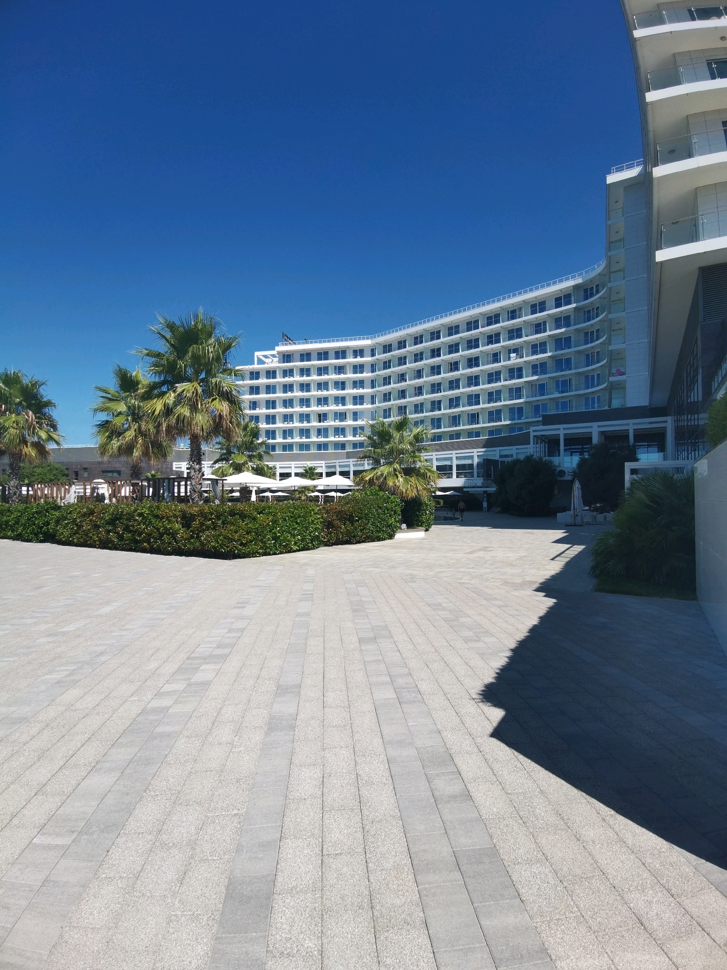 The image shows a large, modern, white hotel building with a curved facade and many windows, set against a clear blue sky. In front of the hotel is a paved courtyard area with a shadow cast by an unseen object or structure. There is a well-maintained hedge and several palm trees between the courtyard and the hotel. The hotel appears to be located in a sunny, outdoor environment, likely a resort or beachfront property. The city of Sirius, Russia is a resort city on the Black Sea coast, known for its modern architecture and tourist attractions. This hotel building is consistent with the type of architecture found in Sirius.
