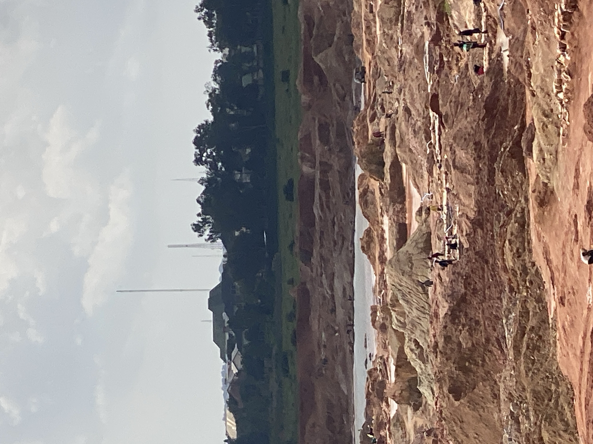 The image shows a group of people engaged in what appears to be rock climbing or rappelling on a steep, rocky cliff face. The cliff is composed of reddish-brown rock, with some lighter-colored sections. There are visible ropes and safety equipment attached to the climbers, indicating an organized activity. Several individuals are positioned at various heights on the cliff, some actively climbing and others seemingly resting or observing. The terrain below the cliff is not entirely clear, but it appears to be a mix of vegetation and possibly water or a dry riverbed in the distance.
To the left of the rocky cliff, there is a distinct change in scenery, with a strip of green grass and dark trees. Beyond the trees, there are some buildings and tall, slender structures that resemble industrial chimneys or towers. The sky is overcast with some diffused light, suggesting it could be daytime, possibly late morning or afternoon. The overall mood is one of adventure and physical exertion, with the people focused on the task of ascending or descending the rock face. The location context provided indicates Reifild-Ist, Nigeria, which might suggest a tropical or subtropical environment given the vegetation. No visible text is present in the image.