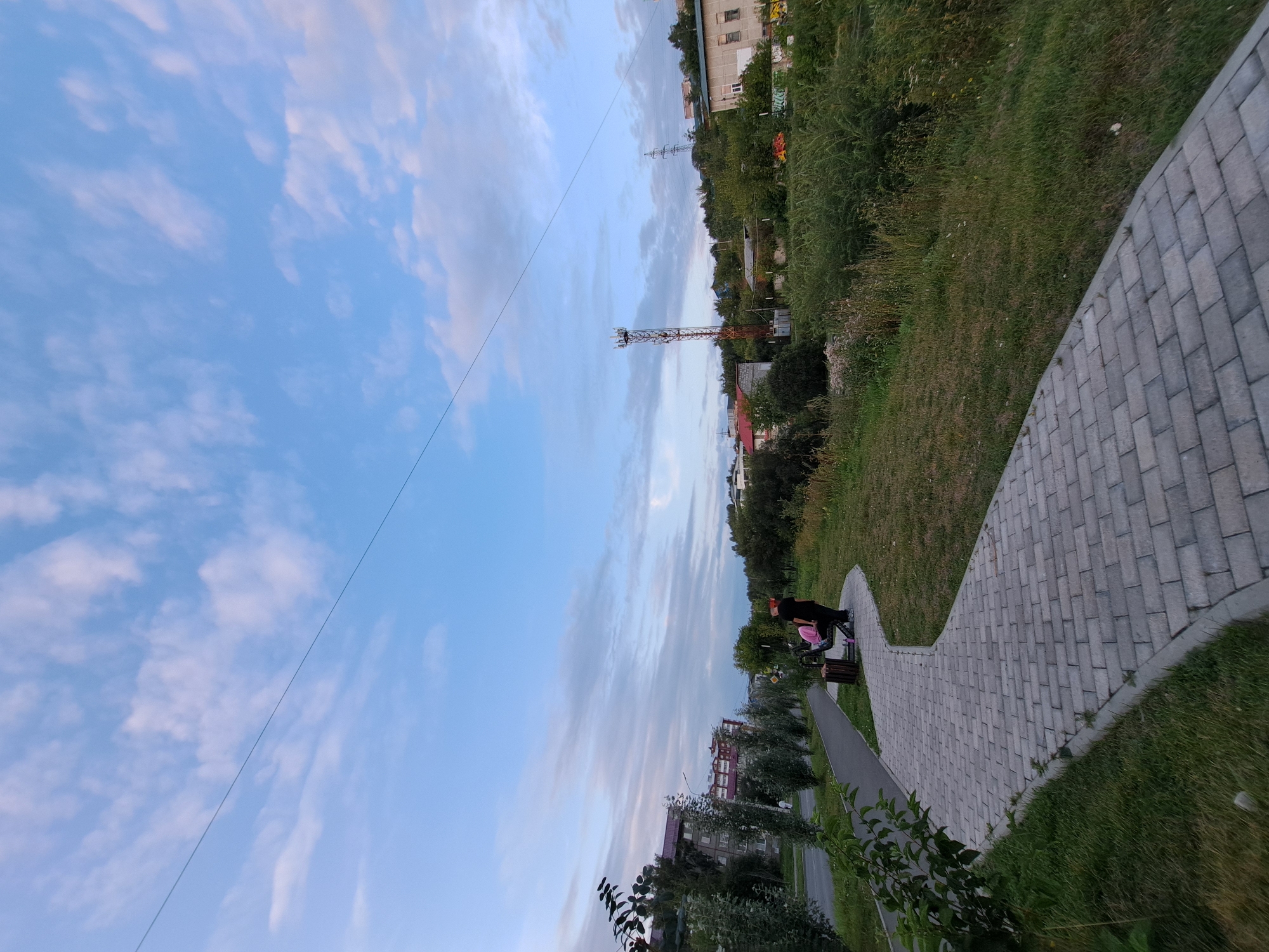 The image captures a view of a city park with a paved pathway winding through it. The pathway, made of light grey interlocking bricks, curves from the foreground towards the left. On the right side of the pathway, a grassy area slopes upwards and is covered with dense vegetation and some taller trees. In the background, a tall, reddish-orange telecommunications tower stands prominently, with several other buildings and some overhead power lines visible. A few people are seated on a park bench near the pathway, one of them wearing a pink garment. The sky above is a soft blue with scattered white and pinkish clouds, suggesting it might be late afternoon or early evening. The scene appears to be in an urban or suburban setting.