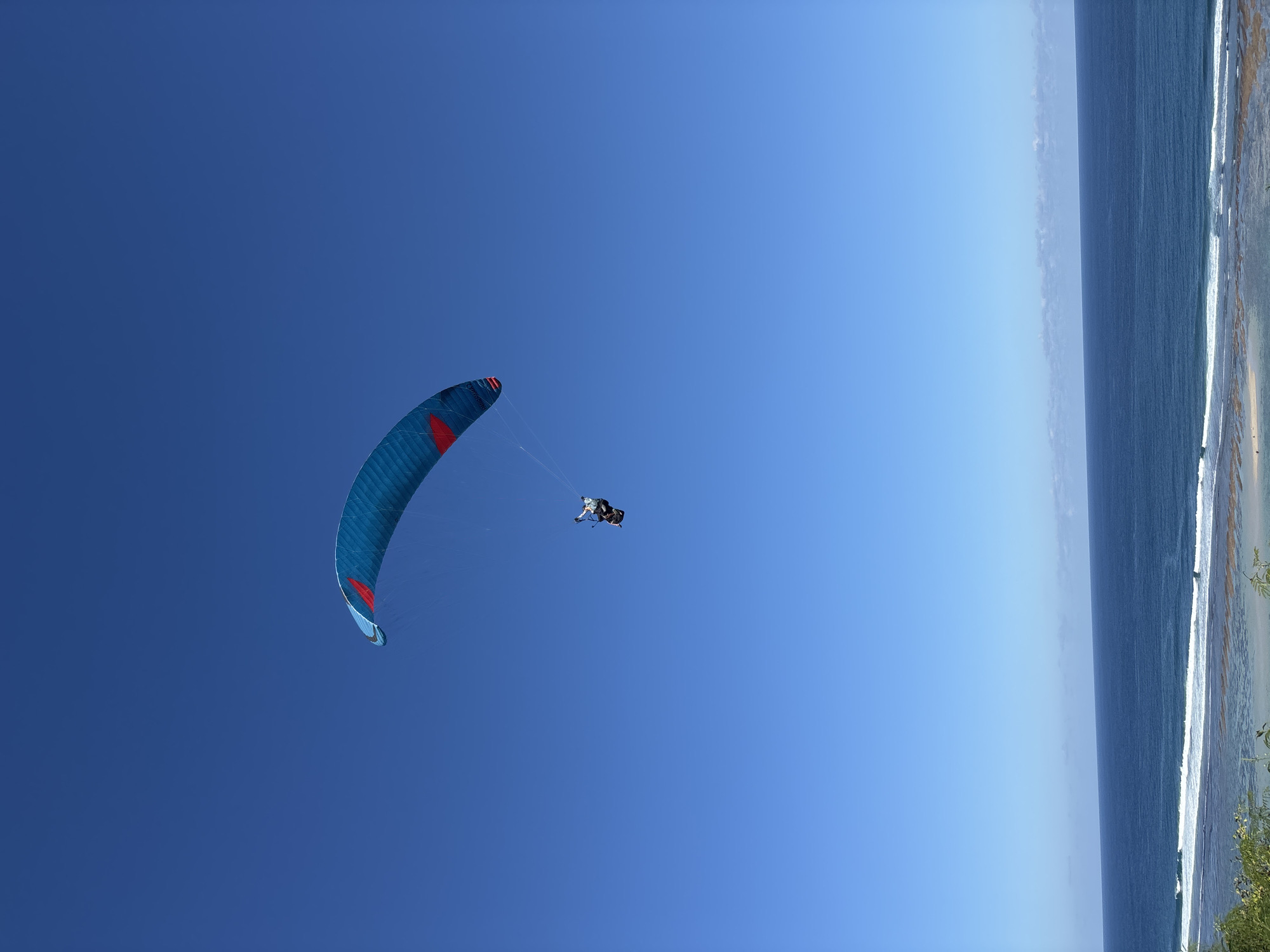 A paraglider soars above the coast of Kuta Selatan, Indonesia. The blue and red paragliding wing is positioned in the left-center of the frame against a clear, deep blue sky. A person is suspended beneath the wing. To the right, the ocean stretches out to the horizon, with breaking waves visible along the shoreline. Vegetation is seen on the coast.