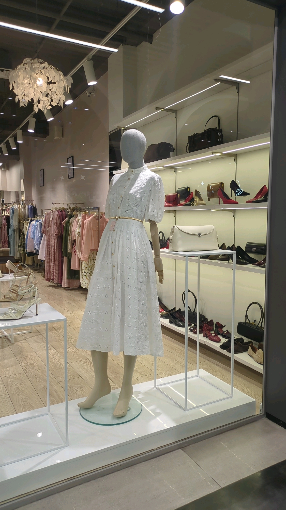 The image captures a storefront display of a clothing boutique. A mannequin, displaying a white broderie anglaise dress with a gold belt, stands prominently in the foreground. Behind the mannequin, shelves are stocked with various handbags and shoes, including red pumps, black heels, and tan loafers. To the left, racks of women's clothing in pastel shades are visible, suggesting a fashion store. The display is illuminated by overhead lighting and accent lights on the shelves, creating a well-lit presentation. The flooring appears to be light-colored wood, and the overall environment suggests an indoor retail space. The reflection in the glass display hints at the presence of a street or mall outside. The location is identified as Chișinău, Moldova.