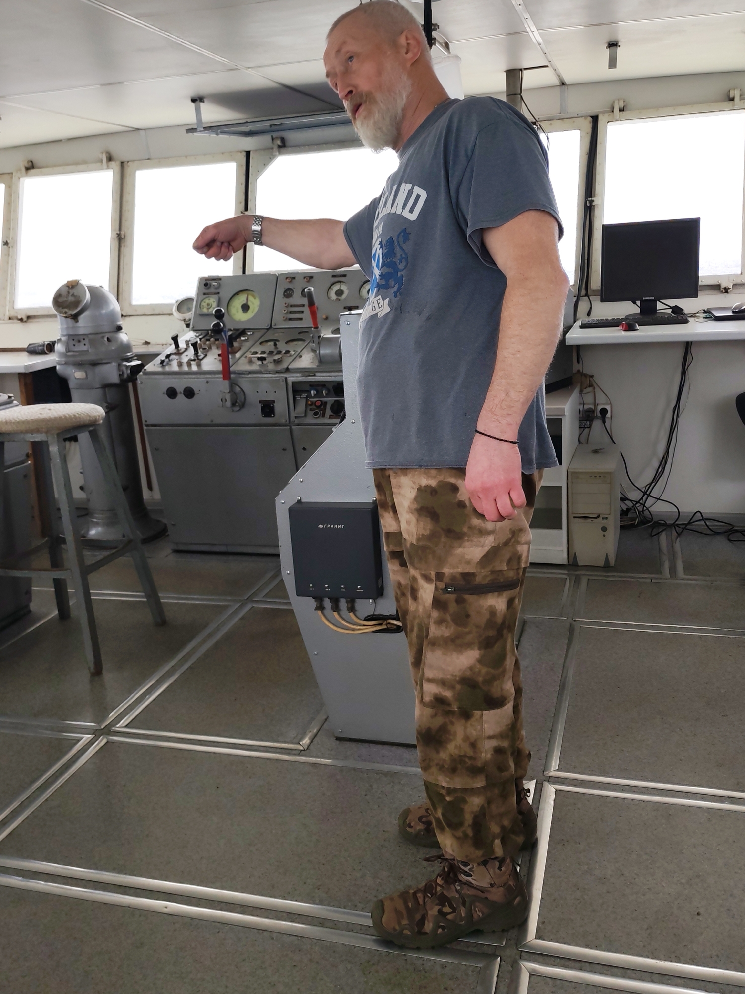 A bearded man, likely middle-aged or older, stands on the bridge or control room of a vessel, gesturing with his left hand. He has a white/grey beard and short hair, wearing a grey t-shirt with "SCOTLAND" and a lion rampant crest, along with camouflage-patterned cargo pants and tactical-style shoes. A silver watch is on his left wrist, and a black bracelet is on his right.
He stands next to a grey control panel equipped with multiple dials, gauges (including one with a yellow face), red and black levers, and various buttons, suggesting navigation or operational controls. To the left of this panel is a large grey stand, possibly housing a gyrocompass, and a simple stool with a light-colored seat. On a console to the man's right, a black electronic device labeled "ΓРАНИТ" (GRANIT) in Cyrillic is mounted, with cables connected at the bottom. Further right, a computer setup comprising a black "SAMSUNG" monitor, keyboard, mouse, and a beige desktop tower sits on a white desk.
The background features multiple rectangular windows revealing a bright, featureless white view, suggesting an overcast sky or foggy conditions outside. The ceiling is light-colored with visible structural elements. The floor has a speckled grey surface with a grid pattern of silver metal strips. The man appears to be explaining or pointing to something within this functional, industrial setting, which aligns with the location context of Shcheleiki, Russia.