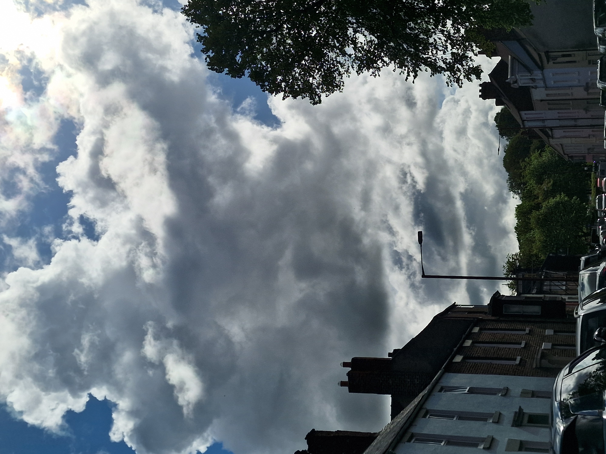 The image shows a view of a cloudy sky above a residential street. The sky is filled with large, gray and white cumulonimbus clouds, with patches of blue sky visible in the lower left corner. In the upper center of the frame, the silhouette of a tree's foliage is visible.
Below the sky, the tops of buildings can be seen. These are traditional brick buildings with pitched roofs and chimney stacks. A street lamp extends from the building. Cars are parked along the side of the street.
The location is likely Durham, United Kingdom, based on the architectural style of the buildings and the general environment.