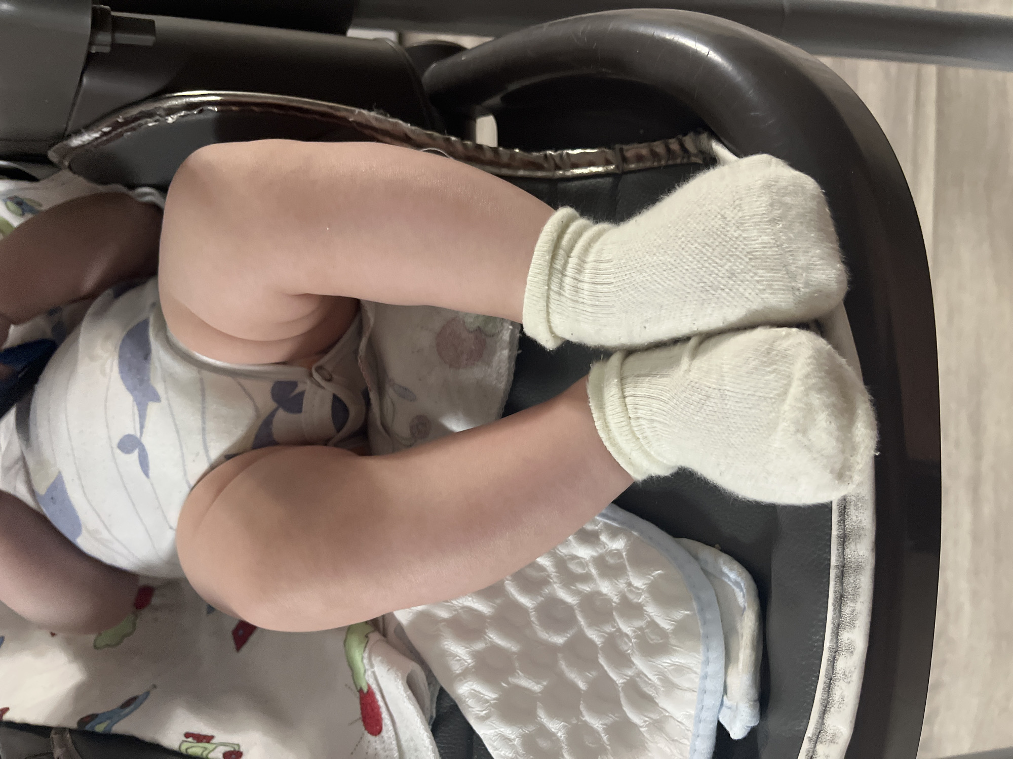 A baby lies on their back in a dark-colored baby swing. The baby is wearing white socks. A disposable diaper can be seen under the baby. The location is unspecified and there is no indication of Раздолье, Russia.