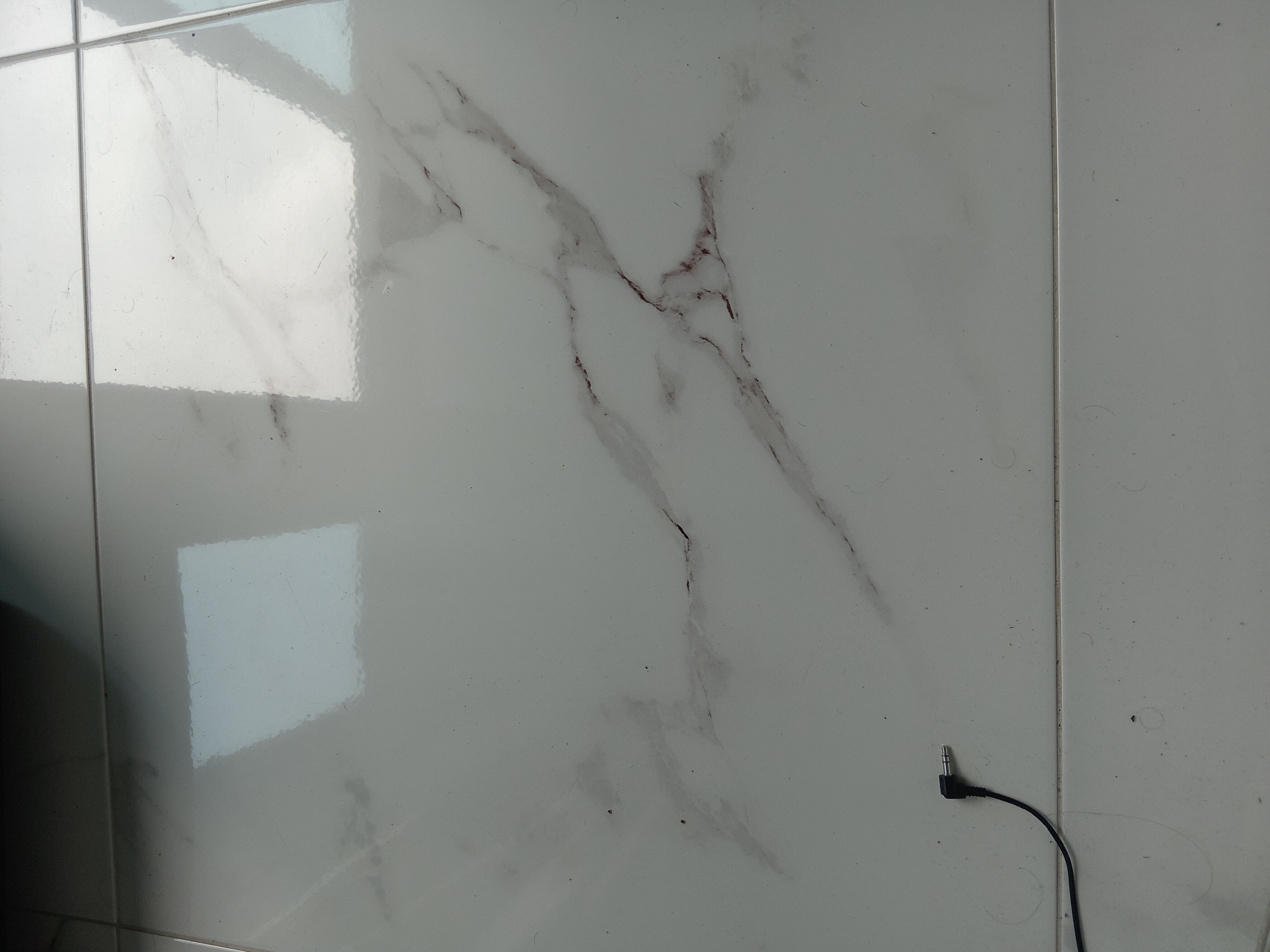 The image shows a section of a tiled wall, likely indoors, consisting of large, light-colored tiles with marble-like veining patterns. A dark-colored audio cable with a 3.5mm connector is visible at the lower right of the frame. The cable runs along the wall surface. The tiles have reflective surfaces showing window reflections. No people are present. The location is unknown, and there is no indication it is located in Batumi, Georgia.