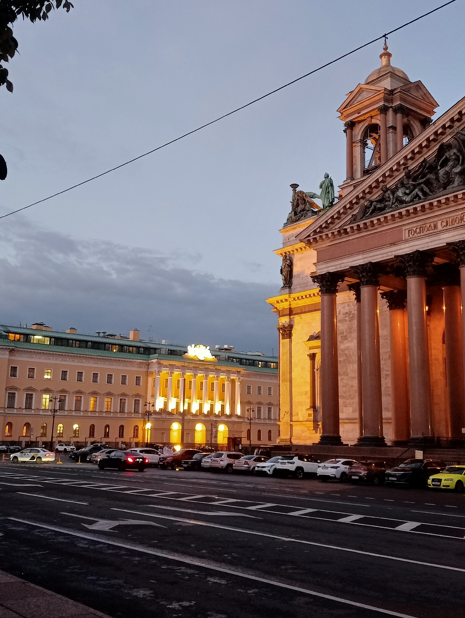 The image captures a view of St. Isaac's Cathedral in Saint Petersburg, Russia, during twilight. The imposing cathedral, with its classical architecture and illuminated colonnade, dominates the right side of the frame. A belfry, also lit, rises above the facade. To the left of the cathedral, a long, pale yellow building with multiple windows, also illuminated, stretches across the background. The sky above is overcast and a dull gray. In the foreground, a street with lane markings is visible, and several cars are parked along the side of the road, facing the cathedral. The overall atmosphere is calm and serene, with the warm light of the buildings contrasting with the cool tones of the sky.