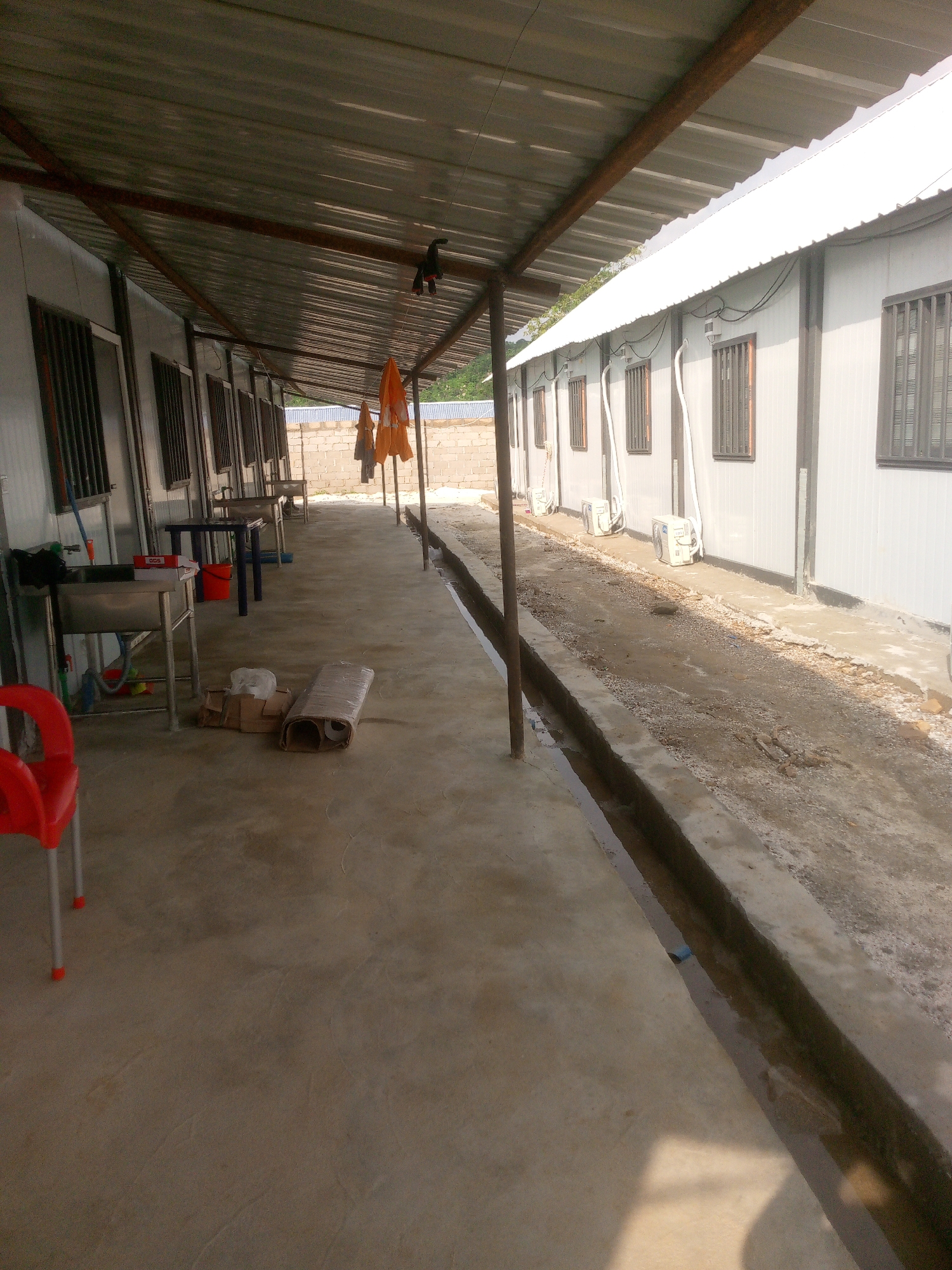The image depicts an outdoor scene in Indo, Nigeria, likely a compound or workers' quarters. The foreground shows a concrete walkway extending to the left, leading towards a series of modular buildings constructed with corrugated metal walls and roofs. These buildings appear to be single-story units, each fitted with barred windows and air conditioning units on the exterior.
On the walkway, to the left, are several metal sinks with plumbing, a red plastic chair, and various items like boxes and rolled-up materials. A bright orange tarp or piece of fabric is visible hanging from a support structure above the walkway, along with what appears to be a black object clipped to a wire. Further down the walkway, more sinks, tables, and orange tarps are visible.
The buildings are situated alongside a narrow, partially filled drainage channel that runs along the edge of the walkway. The area between the drainage channel and the buildings is a dirt and gravel path. The background reveals a brick wall and some vegetation, suggesting the compound is in a semi-urban or rural setting within Indo. The overall impression is one of a functional, possibly temporary, living or working space.