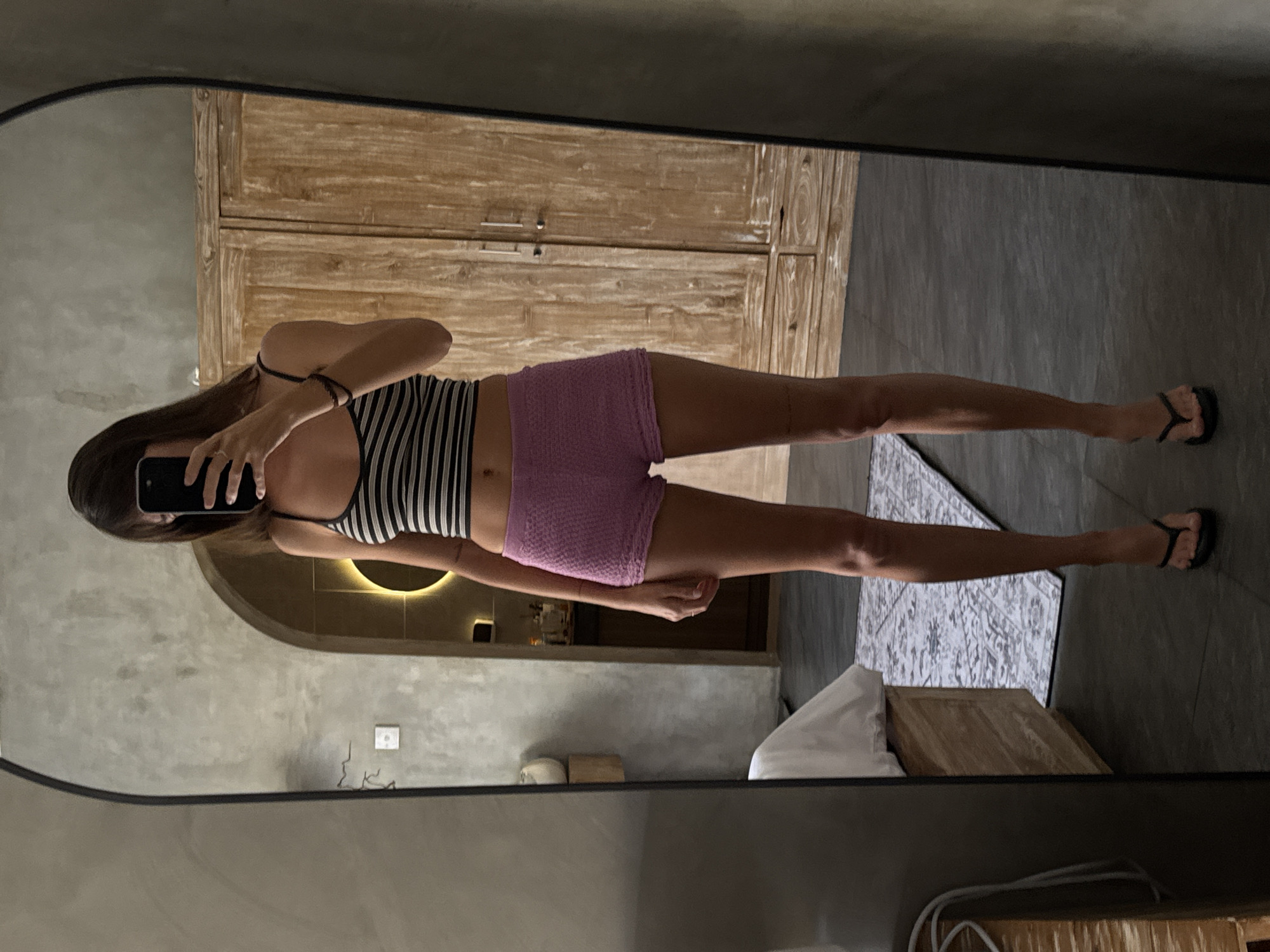 A woman is reflected in a mirror, taking a photo of herself. She is wearing a striped tank top and purple shorts. She is standing on a dark tile floor, and there is a rug visible behind her. In the background, there is a wooden cabinet, a bed with white linens, and a partial view of a room interior with a rounded archway and recessed lighting. The location is in North Kuta, Indonesia.