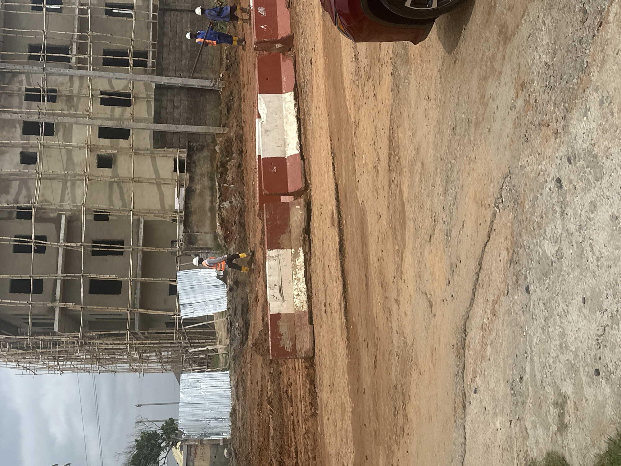 In Gwarinpa, Nigeria, this image captures an active construction site. A multi-story building under construction is visible, enveloped in scaffolding made of bamboo. The building's facade is light-colored, with dark, rectangular window openings. In the foreground, a dirt road is present, marked by rows of concrete barriers painted with red and white stripes, separating different areas of the site. Three construction workers are visible. One worker, wearing an orange vest and yellow boots, walks on the dirt road. Further back, two other workers, one in a blue uniform and another in an orange vest and blue pants, are also present on the construction site. The sky above is overcast.