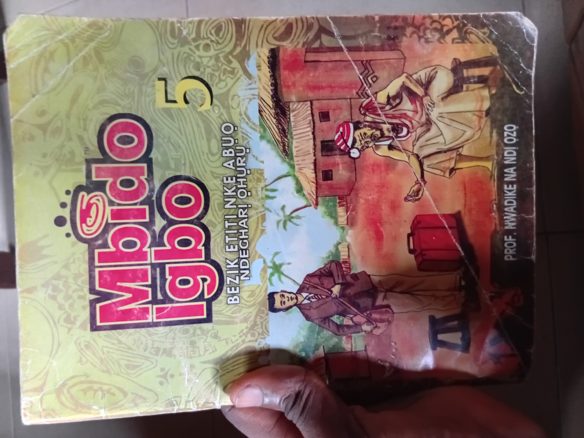 The image displays the front cover of a book titled "Mbido Igbo 5". The book has a light yellow background with decorative patterns. The title "Mbido Igbo" is prominently featured in large, bold, pink letters with black outlines. To the right of the title, the number "5" is displayed in a yellow font. Below the title, smaller text reads "BEZIK ETITI NKE ABUO NDEGĦARI ỌHURỤ".
The illustration on the cover depicts a scene with two figures and traditional buildings. On the left, a man in a brown suit, white shirt, and red and white striped tie stands holding a brown satchel over his shoulder. He is walking towards the right side of the image. In the center and to the right, an older man dressed in a white garment and a red and white striped cap is seated on a stool. He appears to be holding a small object in his left hand. Behind the figures, there are traditional thatched-roof buildings.
At the bottom of the cover, text identifies the author as "PROF. NWADIKE NA NDI OZO".
The image is a photograph of this book cover, held by a person's hand on the left side. The book appears to be old, with some creases and visible plastic covering. The environment depicted in the illustration suggests a rural or traditional setting, possibly in Nigeria, given the Igbo language used in the title and author's name. There is no information in the image that specifically places the scene in Nkpor, Nigeria; the book title refers to the Igbo language and culture, which is prevalent in southeastern Nigeria, including areas around Nkpor.