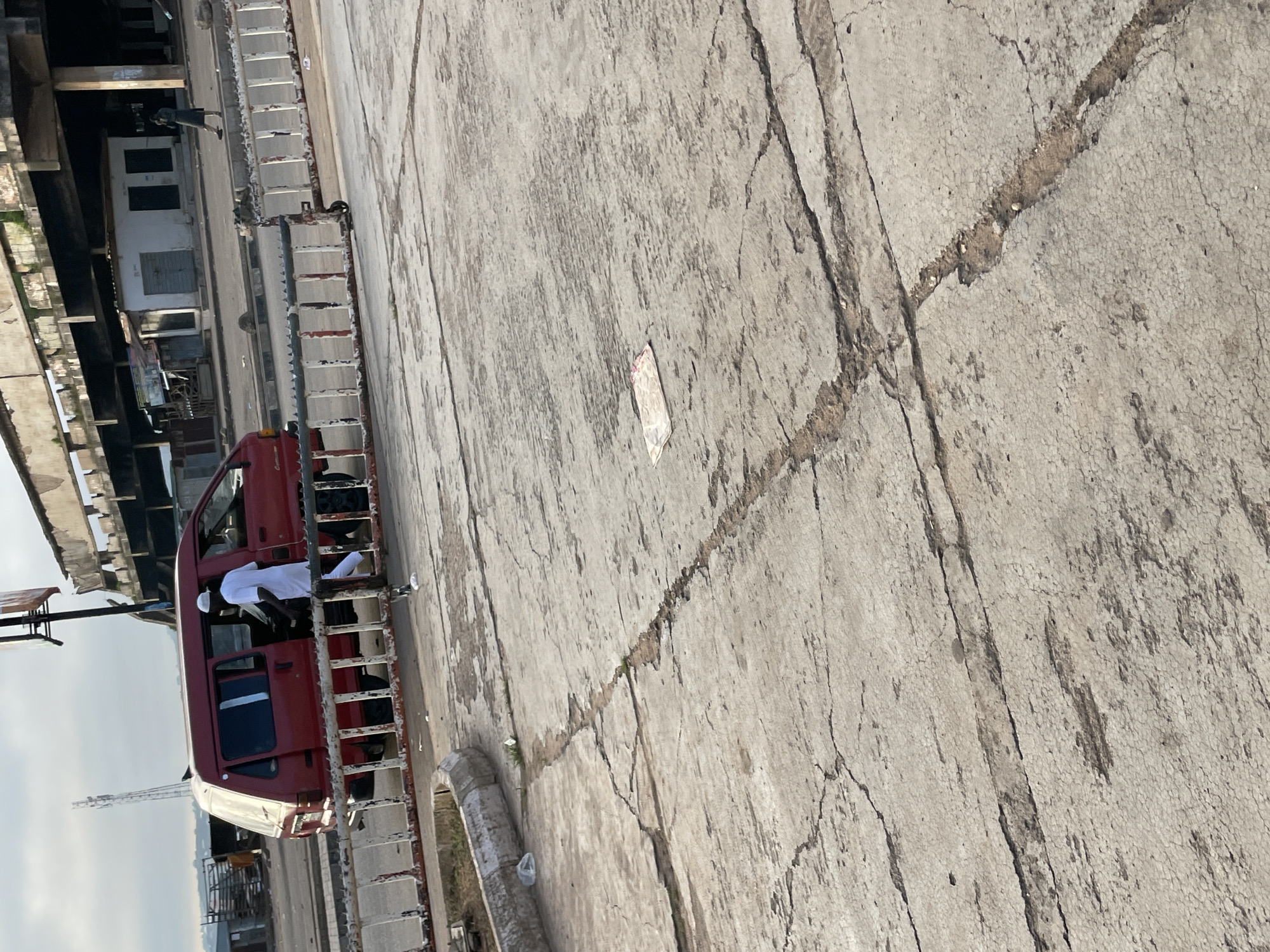 In Jos, Nigeria, a red van is parked next to a metal barrier in an outdoor area with a cracked concrete surface. A man wearing a white robe and cap is exiting the van, with his body turned away from the camera. The van's sliding door is open, revealing the interior. In the background, there is an elevated structure with visible columns and a building with closed doors and windows. A cell tower stands tall in the distance. The ground is littered with debris, including plastic bags and a crumpled piece of paper.