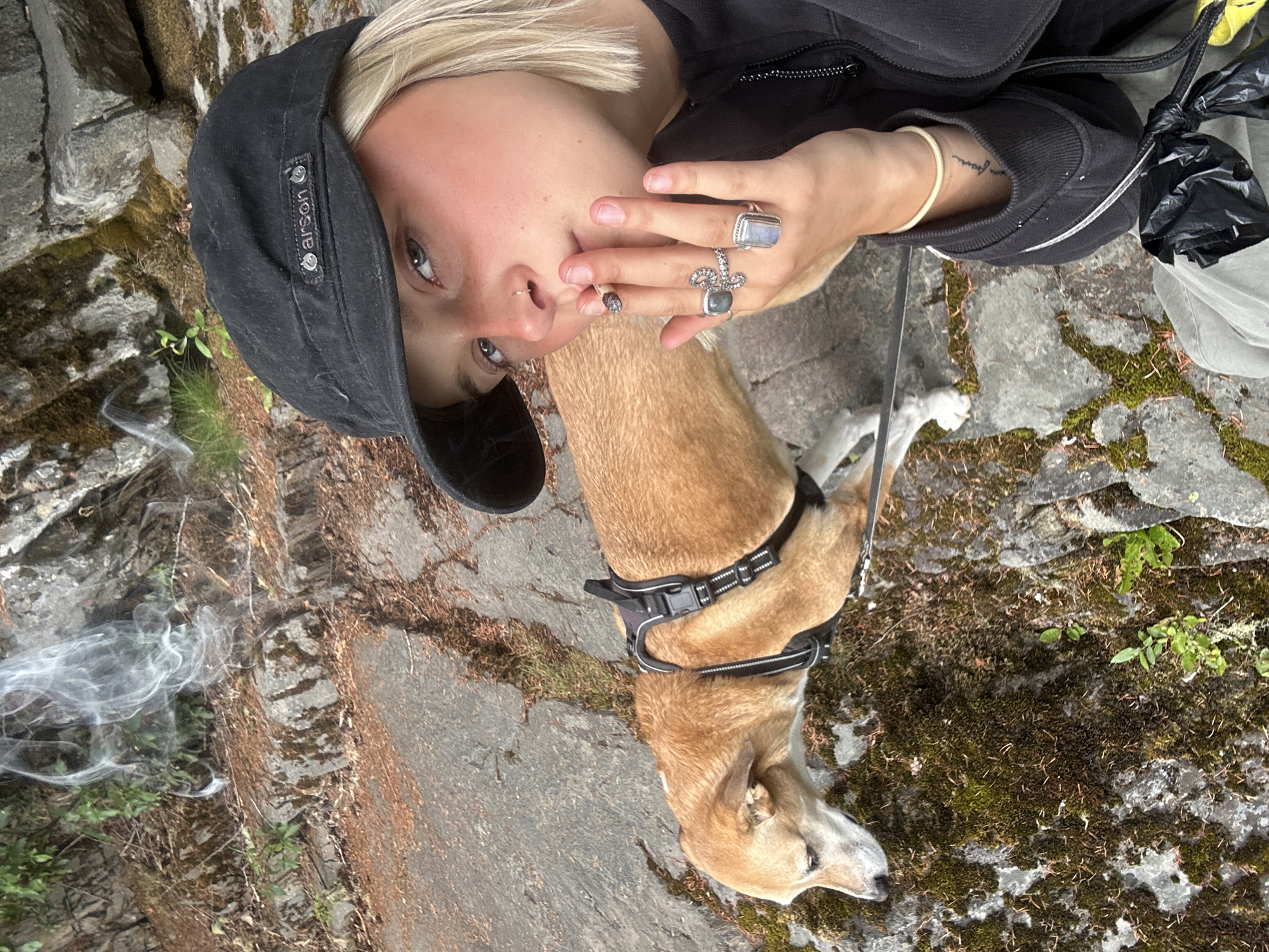 A young woman with blonde hair, wearing a black baseball cap and a black zip-up jacket, is shown from the chest up in the foreground, looking towards the camera with a cigarette in her mouth. She has a nose ring and is wearing multiple rings on her fingers. A light brown dog with a black harness is positioned to her left, looking down and away from the camera. Smoke is visible in the air above the dog. The background consists of rough, rocky terrain with patches of moss and sparse vegetation. The setting appears to be an outdoor natural environment, likely a trail or hillside. There are no visible landmarks that definitively place the location within Whistler, Canada.