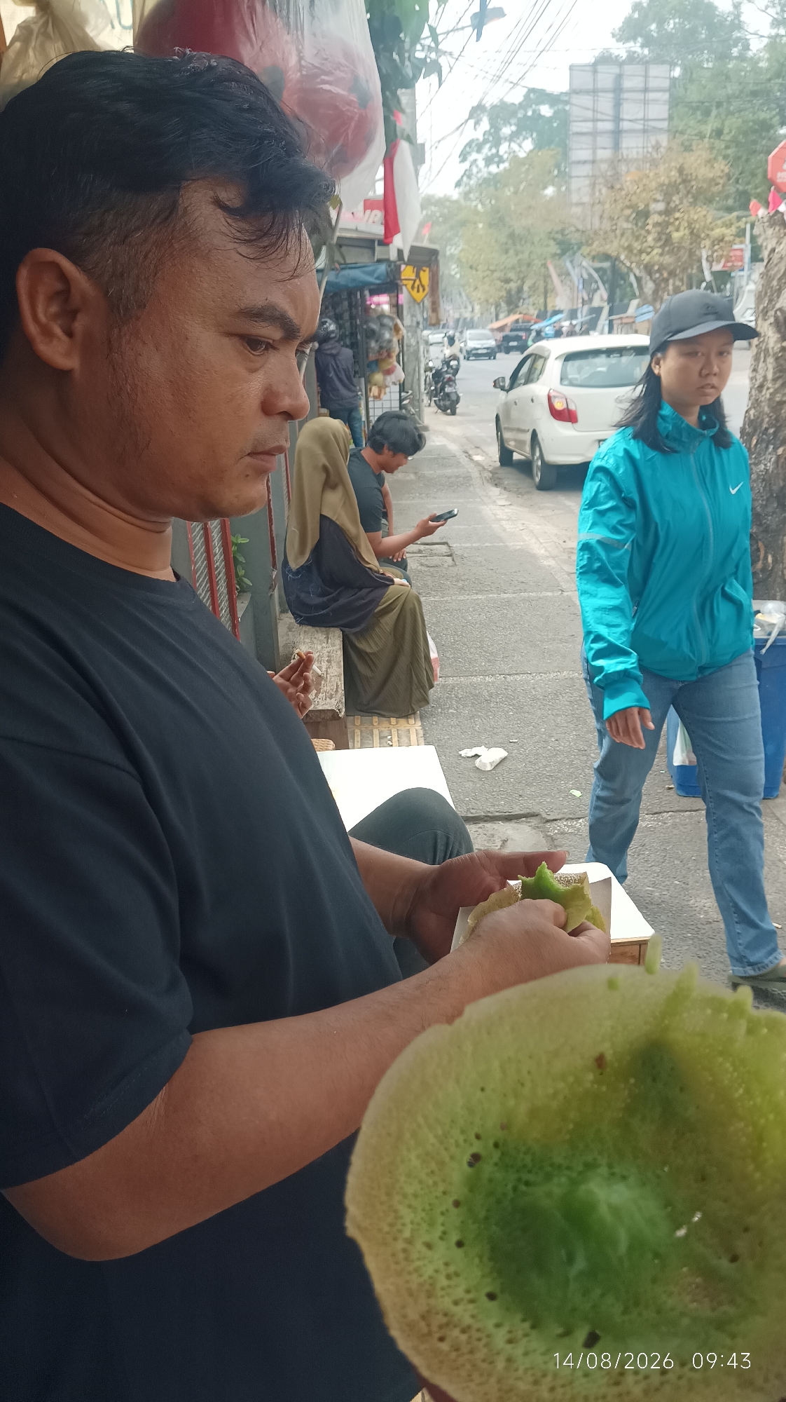 A man in a black t-shirt sits at a white table in the foreground, holding a green pastry and a small piece of the pastry in his hands. He is looking down at the pastry. Behind him, a woman wearing a blue jacket and jeans walks past on a sidewalk. Further in the background, a person in a brown hijab and green dress sits on a bench, looking at a phone. Several cars and motorcycles are visible on the street. The scene appears to be a street-side establishment in Tasikmalaya, Indonesia.