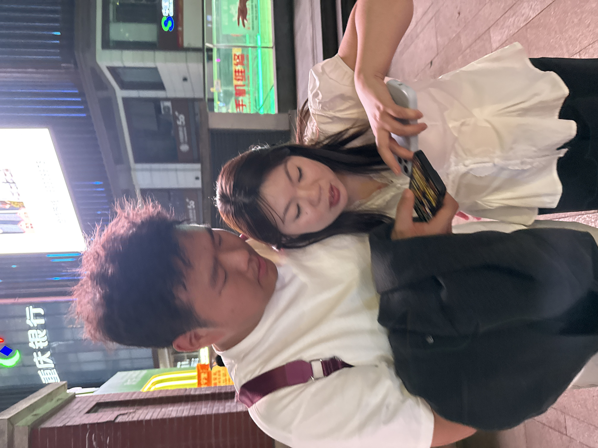 The image captures two individuals, a man and a woman, interacting with a smartphone in an urban setting at night. The scene is brightly lit by numerous artificial lights and neon signs from surrounding commercial buildings, indicating a bustling city environment.
The woman, positioned on the right side of the frame, has long dark hair and wears a white, ruffled blouse. She leans towards the man, her gaze directed at the smartphone he holds, with her mouth slightly open as if speaking or reacting. She is also holding a light-colored smartphone in her left hand.
The man, on the left, has short, somewhat messy dark hair and is wearing a white long-sleeved shirt. A burgundy strap, possibly from a bag or an accessory, is visible across his chest. He holds a smartphone in his right hand, intently focused on its screen. A dark jacket or bag is draped over his left shoulder.
The background features several building facades with large windows and various illuminated signs. On the far left, a very bright light source, possibly a large LED screen, dominates a portion of the background. The ground appears to be paved.
The primary activity depicted is the shared viewing of a smartphone, suggesting they are looking at content together. The female's expression suggests engagement, possibly amusement or surprise.
Visible text on the signs is in Chinese, consistent with the specified location of Yuzhong District, China. A prominent sign on the left displays "重庆银行" (Chóngqìng Yínháng), which translates to "Chongqing Bank." Other signs in the background show "5G" and include partially decipherable characters that suggest "手机店" (Shǒujī diàn), meaning "mobile phone store," and "手机贴膜" (Shǒujī tiēmó), or "mobile phone screen protector." A green sign also features a hand making a "V" gesture, and a blue "S" logo is visible on another sign.