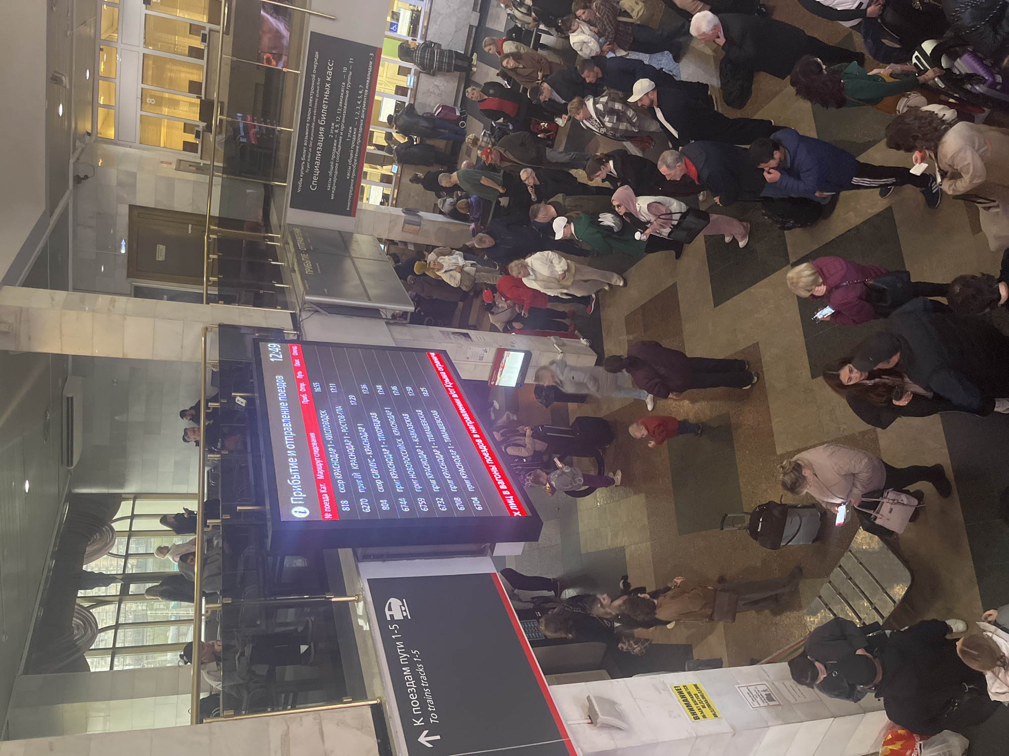The image shows a crowded train station in Krasnodar, Russia. Many people are standing and waiting near a large electronic departure board displaying train schedules and destinations such as Krasnodar to Kislovodsk, Rostov-Glavny, and Timashevskaya. A sign indicates directions to trains on tracks 1-5. Signs with Russian text provide information about ticket office specializations and mention electronic queues. People are waiting near the ticket counters, and some are looking at their phones.