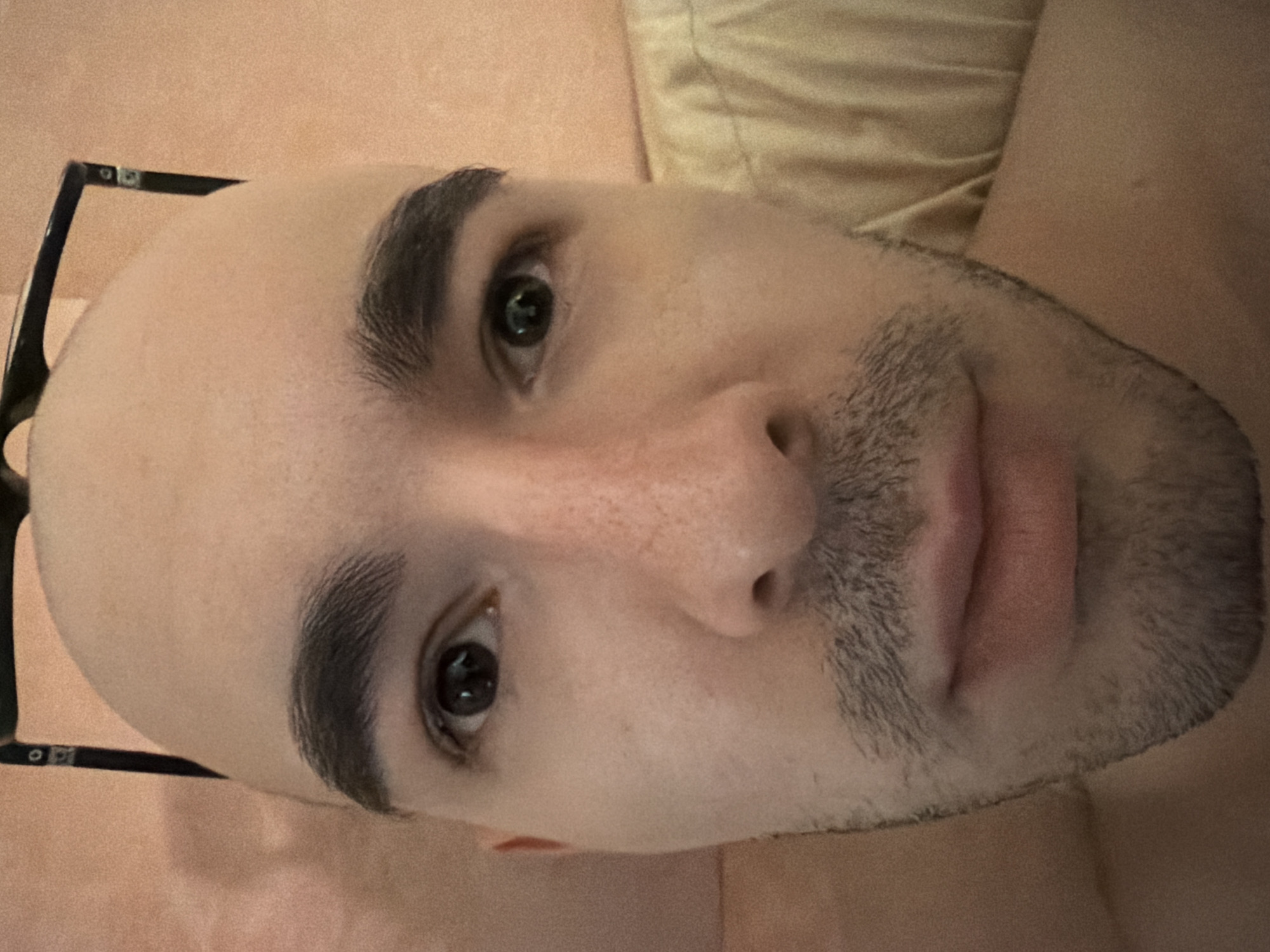 A bald man with thick eyebrows and a stubbly beard lies on a bed, his face angled towards the camera. He is wearing glasses that are partially visible on the left side of the frame. The background includes a pillow. The location is unknown.