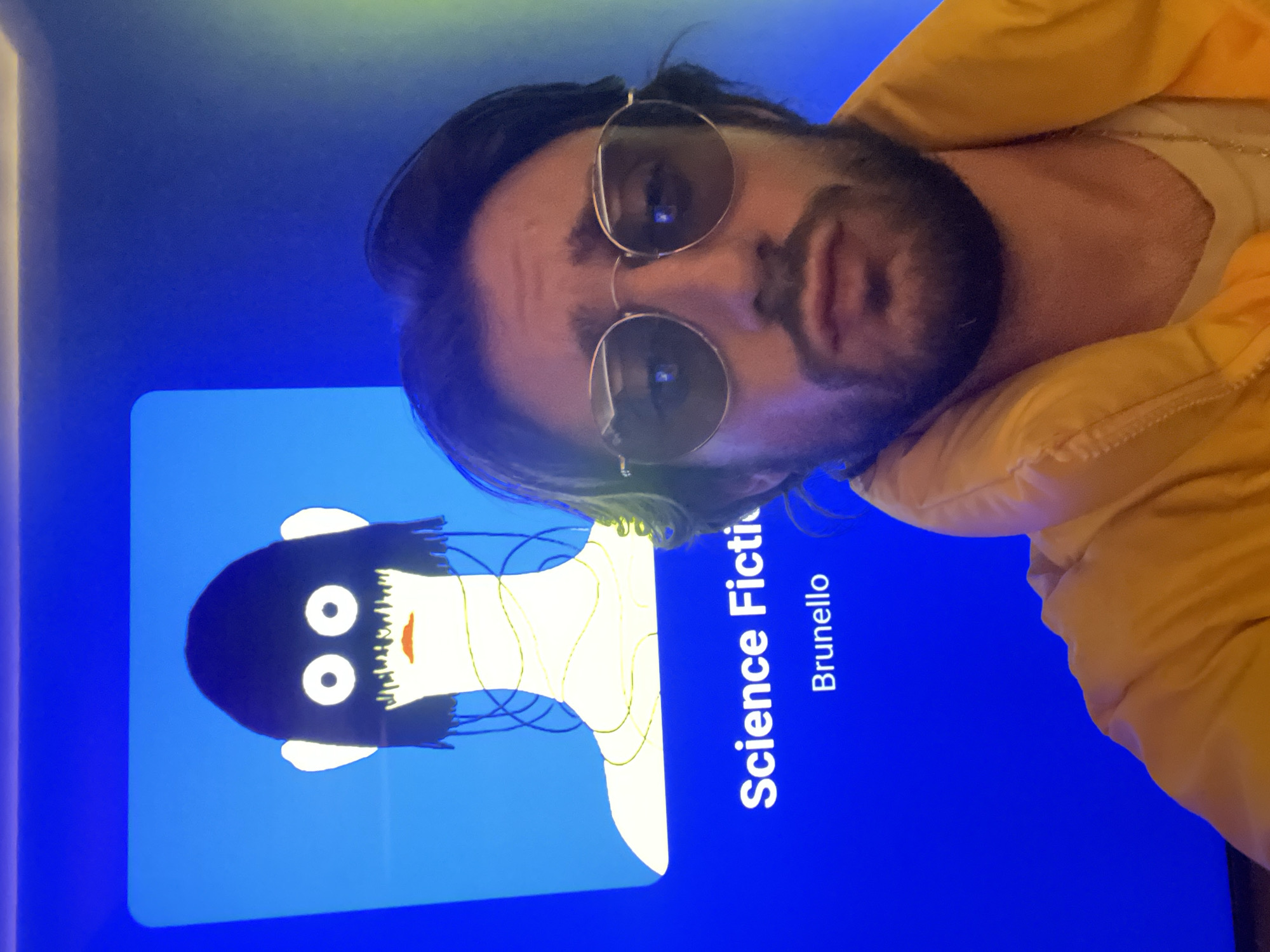 The image shows a man with dark hair, a beard, and sunglasses wearing a yellow jacket. He is positioned in front of a blue screen displaying a cartoonish image of a figure with a black head, white eyes and ears, and a red mouth. The figure is connected by lines to a white body shape. Text on the screen reads "Science Fiction" and "Brunello." The location is not discernible from the image.
