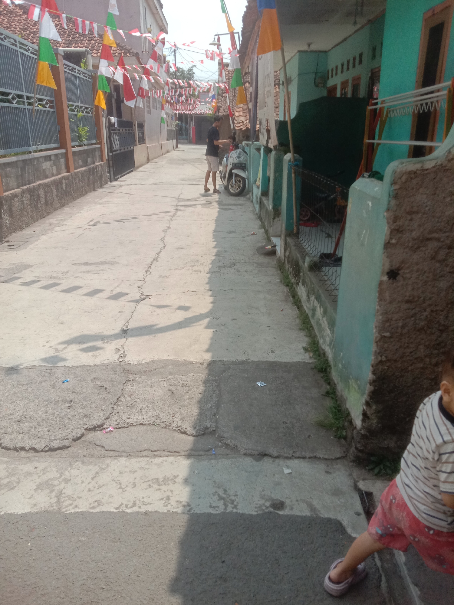 The image captures a narrow street in Bojong, Indonesia, adorned with Indonesian flags and banners, indicating a festive occasion. The street is paved with concrete, showing signs of wear and tear with cracks and uneven surfaces. Houses line both sides of the street, with one house painted in a vibrant teal color. A white scooter is parked on the side of the road, and a clothes drying rack is visible in front of the teal house. A man in shorts and a t-shirt stands near the scooter. In the foreground, a young child wearing a striped t-shirt and red shorts with a pattern is partially visible, walking or standing on the pavement. The sun casts shadows across the street, suggesting it is daytime. The overall scene depicts a typical residential street in Indonesia during a celebration.