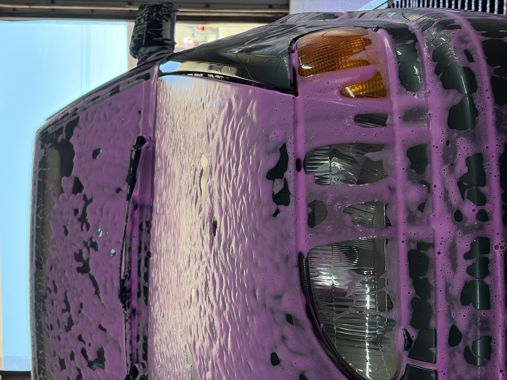 A close-up shot depicts a vehicle undergoing a wash, heavily coated in pink soap suds. The visible features include a headlamp, a turn signal, a portion of the vehicle's door, and a side mirror. The vehicle appears to be in a car wash facility, indicated by the soapy residue and the architectural elements visible through the window, likely the structure of the car wash itself. The specific location in Chaikovsky, Russia, cannot be determined solely from this image.