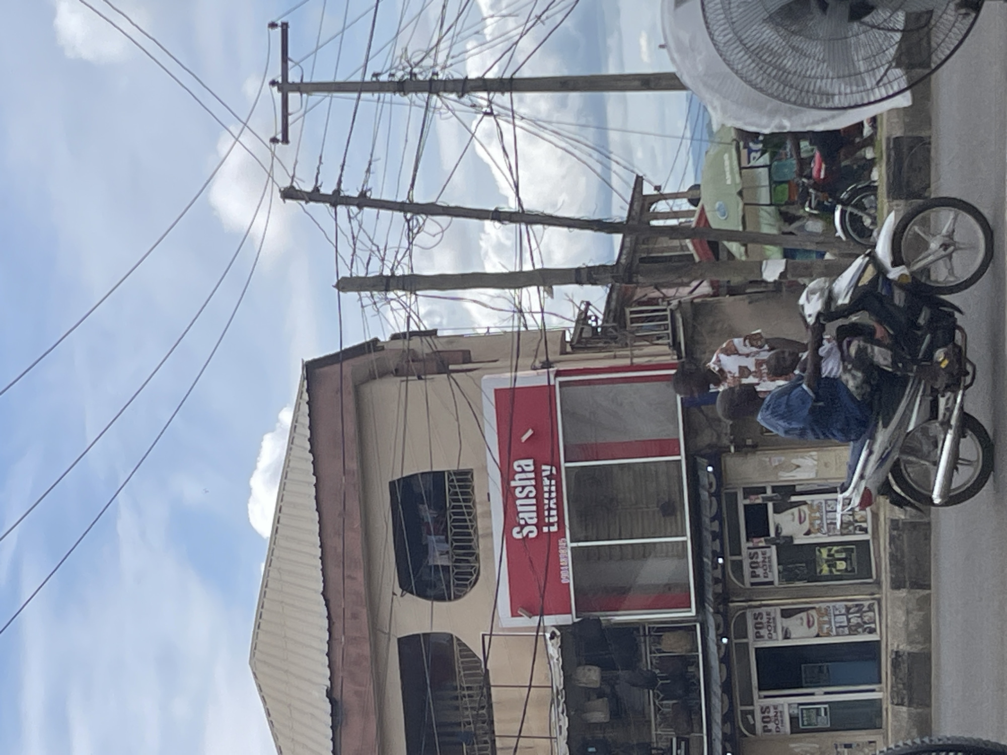 In an outdoor, urban setting, a motorbike carries two people, a man and a child, down a street. The man, wearing a blue shirt, is driving, while the child, in a white shirt, sits behind him. The motorbike, a silver and white model, is in motion. Behind the motorbike, a storefront displays signs that read "POS DONE" and a large image of a woman's face. Above this storefront, a red sign reads "Sansha Luxury." To the right of the storefront, utility poles are laden with numerous wires. In the background, additional buildings and a green umbrella can be seen. The sky is partly cloudy.