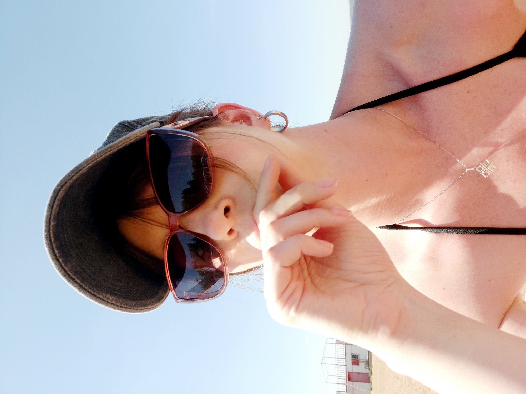 A person wearing sunglasses and a hat is standing outside under a clear blue sky. They are wearing a black bikini top and a necklace with a geometric pendant. Their hand is raised to their face. In the background, there is a building with a red door and a white cage-like structure on top. Based on the presence of the building, the location is Chaikovsky, Russia.