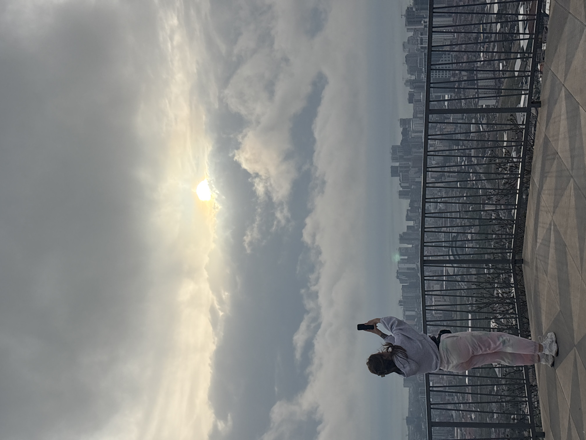 The photo shows a person holding a cell phone and taking a picture of a cloud-filled sky and a distant cityscape. The setting appears to be an elevated observation point with a metal railing and a patterned stone floor. The person is wearing a light-colored sweatshirt and tie-dye sweatpants. The cityscape suggests an urban location, and it's a view from the Alphabetic Tower observation deck in Batumi, Georgia, looking towards the city. The sun is partially obscured by clouds, creating a bright, diffused light in the sky.