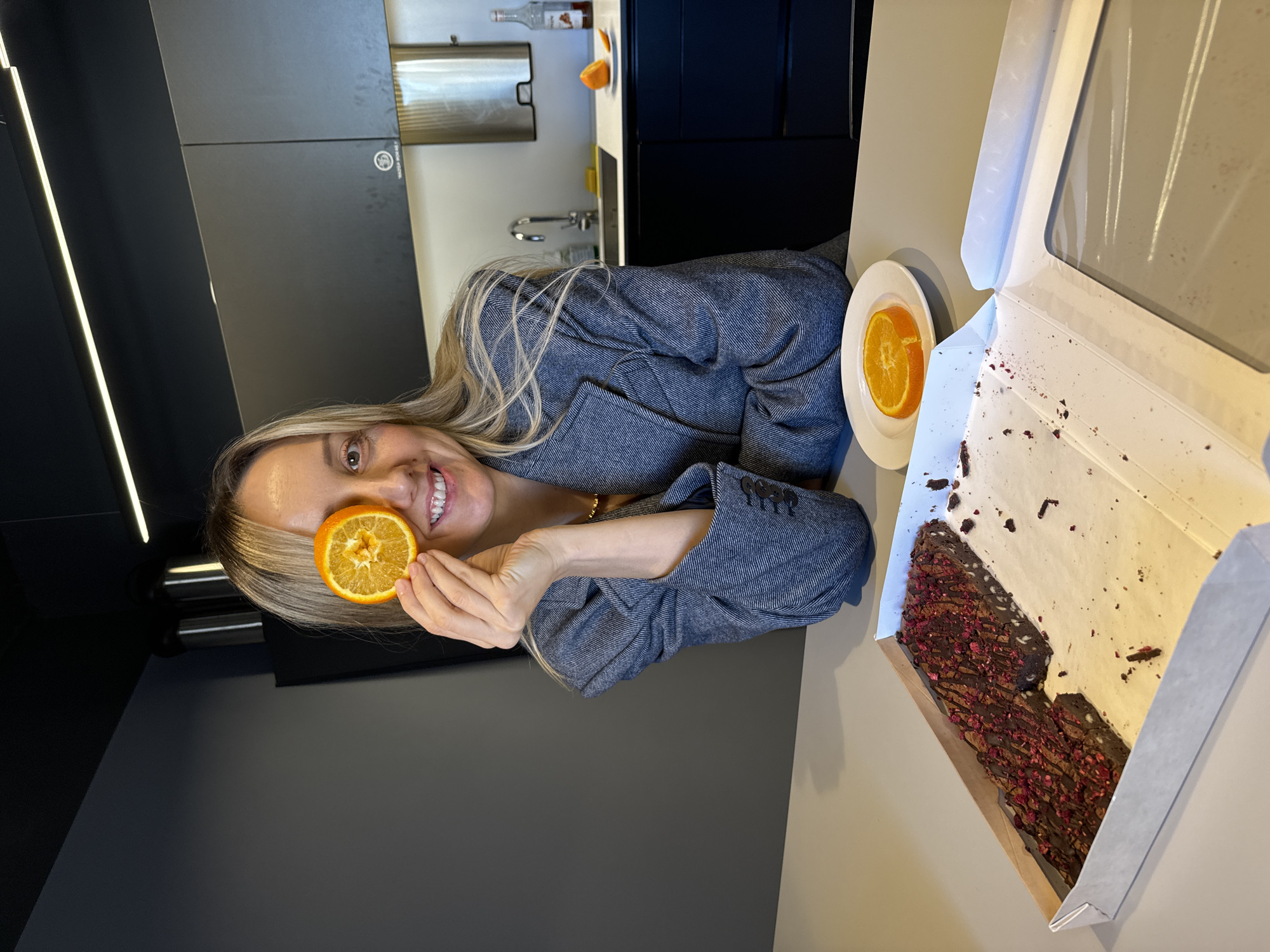 A woman is seated at a table indoors. She is holding a half of an orange in front of her left eye and smiling. She is wearing a grey jacket. On the table, there is a white plate with another half of an orange and an open box with a chocolate dessert that appears to have raspberry toppings. In the background, there is a modern kitchen with dark grey cabinets. A bottle and an orange are on a counter. The location is possibly in Riga, Latvia.