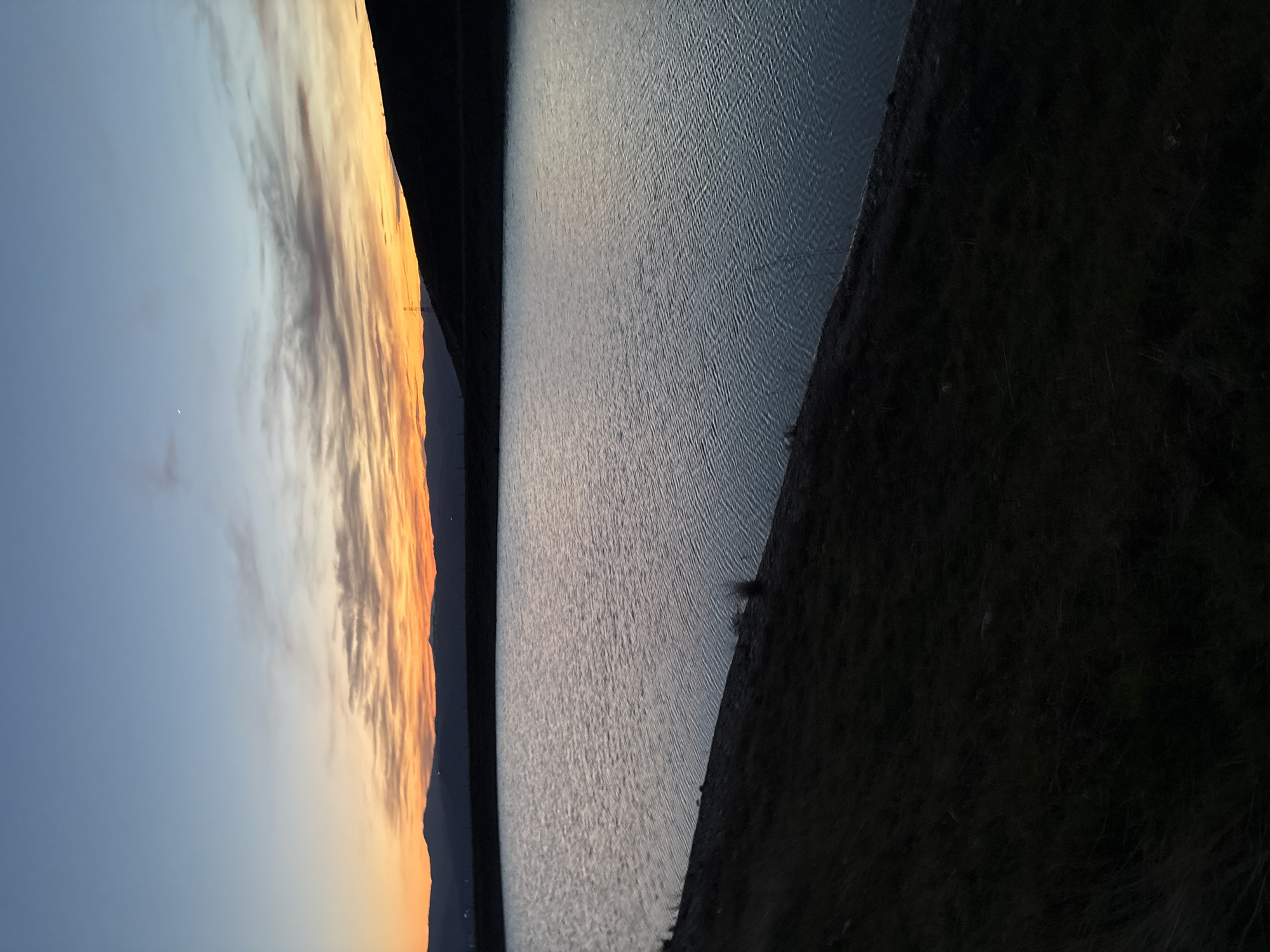 The image captures a landscape scene likely at dusk or dawn. On the left side, a sky transitions from blue to orange and yellow hues, suggesting a sunrise or sunset. Linear clouds stretch across the horizon. Adjacent to the sky is a body of water, its surface reflecting the light of the sky, with small ripples visible. Along the right edge of the water, a dark, steep embankment rises, possibly a hill or mountainside, covered in dark vegetation. The entire scene is shot at an angle, rotated counter-clockwise. The location is mentioned as Polenitsa, Bulgaria. There are no people visible.