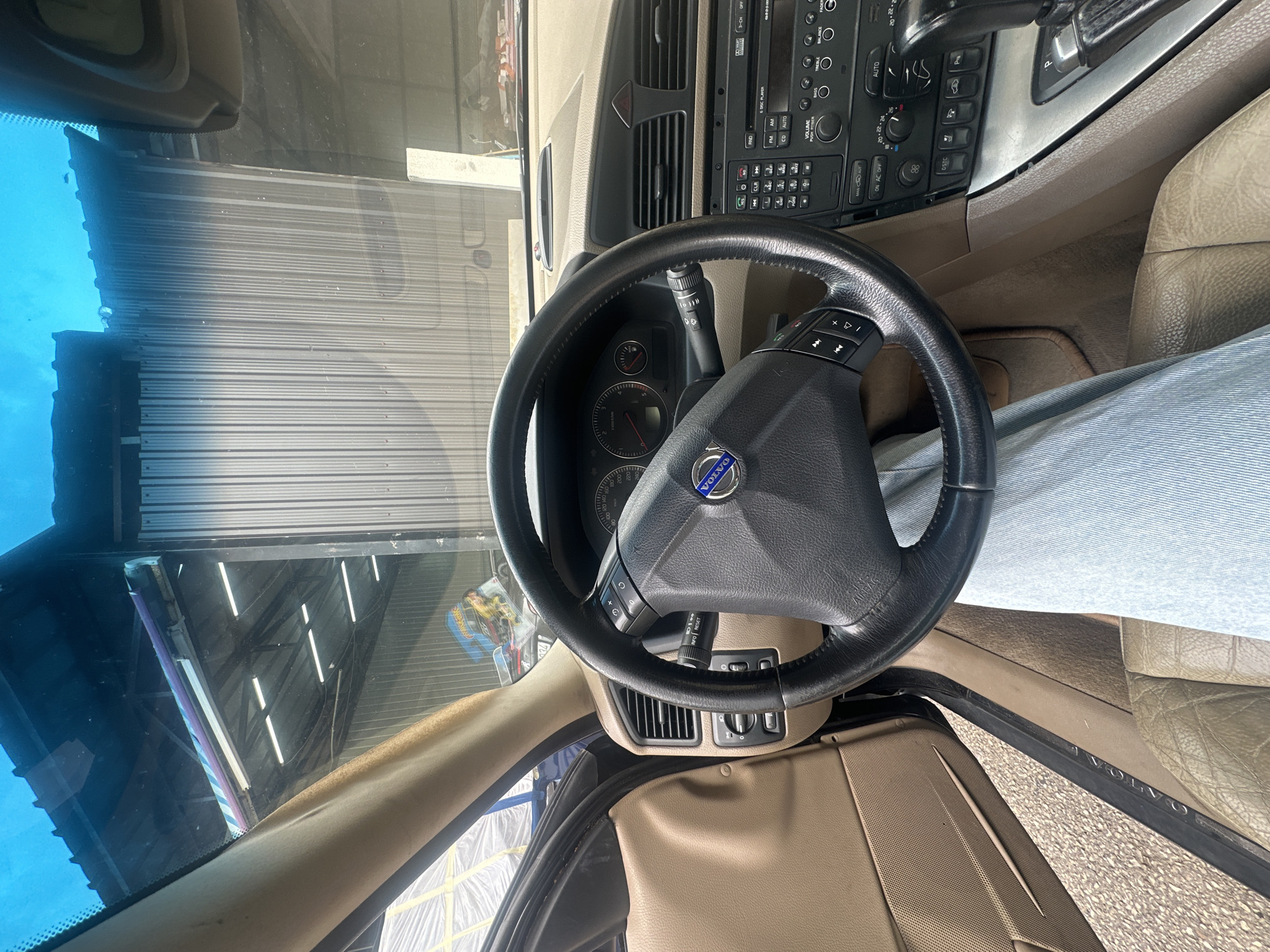 The image displays the interior of a Volvo car, captured from an elevated perspective looking towards the driver's side and dashboard. The photograph is oriented sideways, with the car's front facing to the left.
In the foreground, a black, three-spoke steering wheel prominently features the "VOLVO" emblem in its center. The left spoke of the steering wheel has controls for audio and cruise control. Behind the wheel, the instrument cluster shows a speedometer and tachometer, with numerical markings and indicator lights. A stalk extending from the steering column on the right side bears the text "INFO RESET."
The car's dashboard is a tan or light brown color. The center console houses an older model car stereo and CD player, with clearly visible buttons and text including "FM AM CD AUTO," "VOLUME PUSH POWER," and controls for "BASS TREBLE BALANCE FADE," and "3-CH OFF." Below the stereo, climate control settings display "AUTO," "ON AC OFF," and temperature values like "20," "22," "24," along with an "88" display, likely indicating fan speed or a radio frequency. Other buttons on the console include "DSTC" (Dynamic Stability and Traction Control) and "P" (parking sensor).
A portion of a person's legs, wearing light blue pants or jeans, is visible in the driver's footwell, suggesting the presence of a person in the driver's seat. The car's interior is finished with tan leather seats and matching plastic trim panels. The driver's side door sill also has "VOLVO" embossed on it.
Through the front windshield, a reflection reveals an indoor industrial setting, characterized by corrugated metal walls and bright fluorescent strip lights on a dark ceiling, indicating the vehicle is likely situated in a garage or workshop. A yellow Hot Wheels package is faintly discernible in a reflection near the upper-left corner of the windshield. The interior appears used but well-maintained. The location context is Budva, Montenegro.