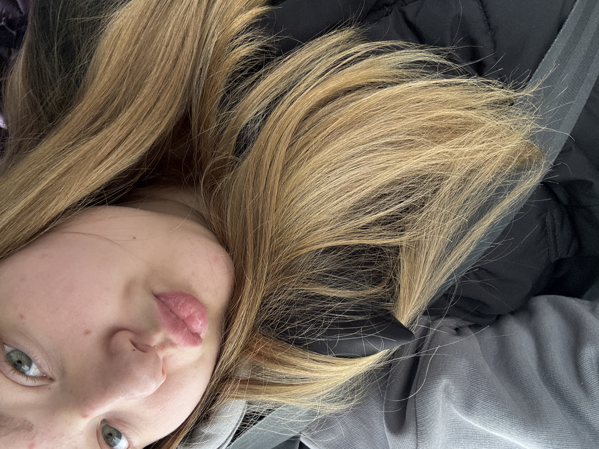 A close-up, rotated image features a young individual with light brown hair spread across the frame. The person has fair skin and light green eyes. The face is partially visible, with the left side of the face and one eye more prominently displayed. The subject's lips are a light pink shade. The background consists of dark clothing, possibly a jacket, and a gray fabric, likely part of a sweater or other garment. There is no information in the image to determine if the image was taken in the city of Chaikovsky, Russia.