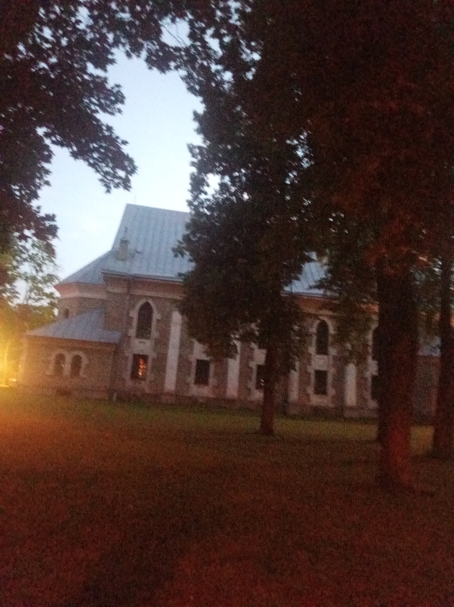 The image shows a view of St. Lawrence's Church in Tapa, Estonia, at dusk. The church is a stone building with a light-colored facade and a gray, sloped roof. It has several arched windows, some of which are illuminated from within. In the foreground, there is a lawn area, and trees with dark foliage partially obscure the view of the church. The sky is a pale, hazy blue, indicating the transition from day to night. The overall lighting suggests a low-light environment, possibly due to artificial lighting from within the church and an external light source on the left side of the image. No people are visible in the scene.