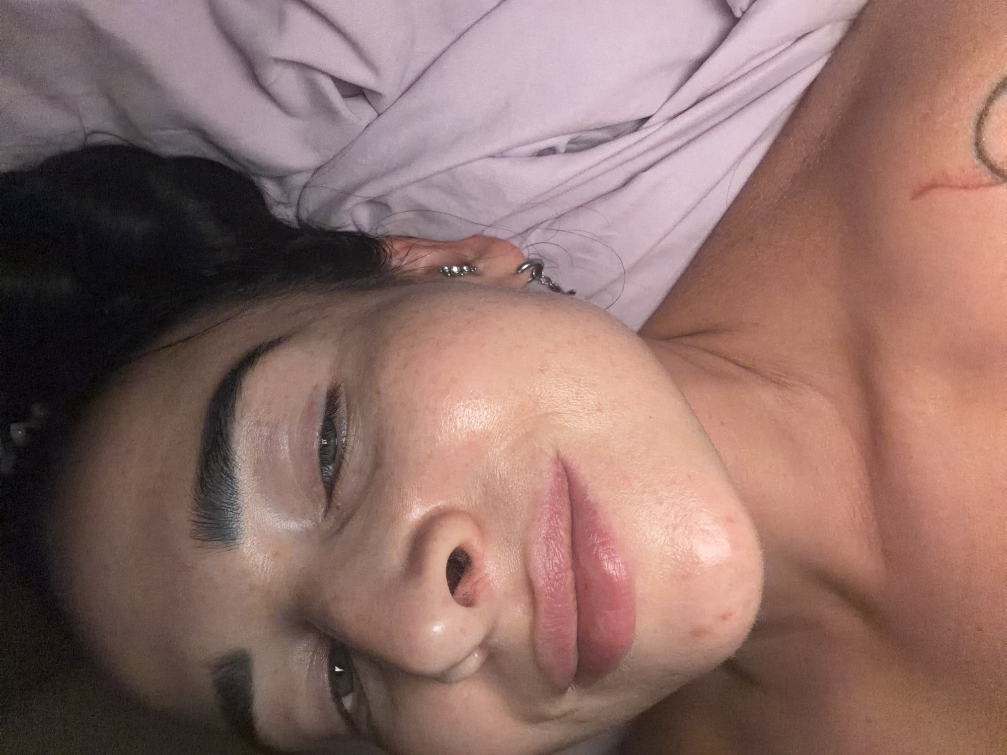 A close-up, high-angle shot captures a person lying down, possibly in bed, with their head resting on a lavender-colored fabric, likely a bedsheet or blanket. The person's face is oriented diagonally, with their right side and part of their torso visible. They have dark hair, visible along the top and side of their head. Their eyes are partially closed, and their eyebrows are dark and well-defined. The lips appear full, and the skin on their face has a somewhat glossy, hydrated appearance.
Notable details include multiple silver-toned piercings on their right ear and a partial view of a circular tattoo design on their right shoulder or upper arm. A reddish mark, possibly a scratch, is also visible near the tattoo. The lighting is soft, suggesting an indoor setting. The person's expression appears calm and relaxed. No specific activity is clearly depicted beyond resting. There is no visible text in the image. While the provided context suggests Saint Petersburg, Russia, there are no visual cues within the image to confirm this location.