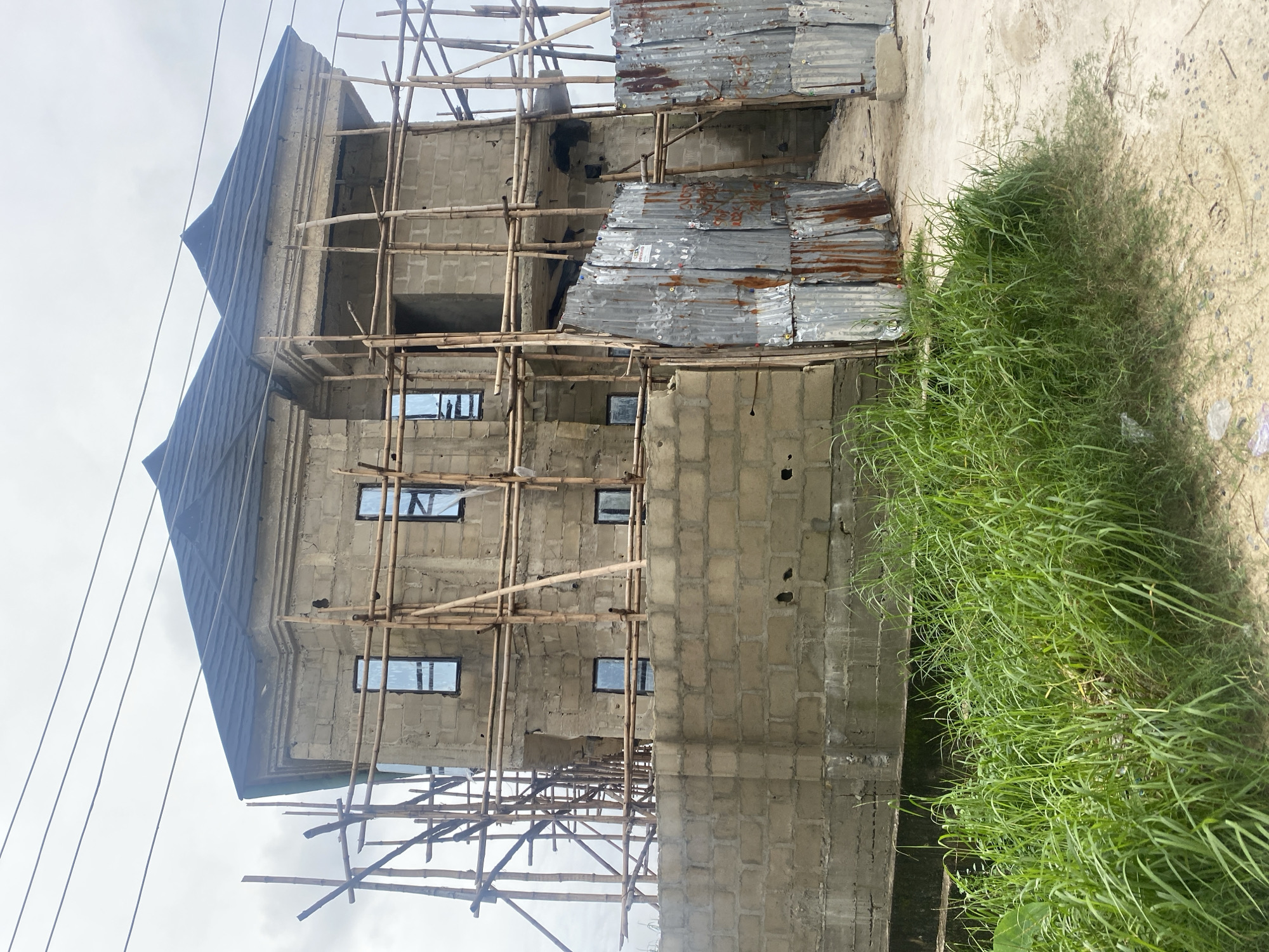 A partially constructed, multi-story building, likely a house, is the central subject of the image, set against an overcast sky. The structure is composed of concrete blocks and is surrounded by bamboo scaffolding, indicating ongoing construction. Several windows are visible, fitted with reflective glass. A pitched, dark-colored roof tops the building. In the foreground, tall green grass lines the bottom of the frame. The entrance area appears to be partially screened by corrugated metal sheets, some of which show signs of rust. The ground surface is unpaved dirt. The overall scene depicts an unfinished residential construction project in what appears to be a developing area.