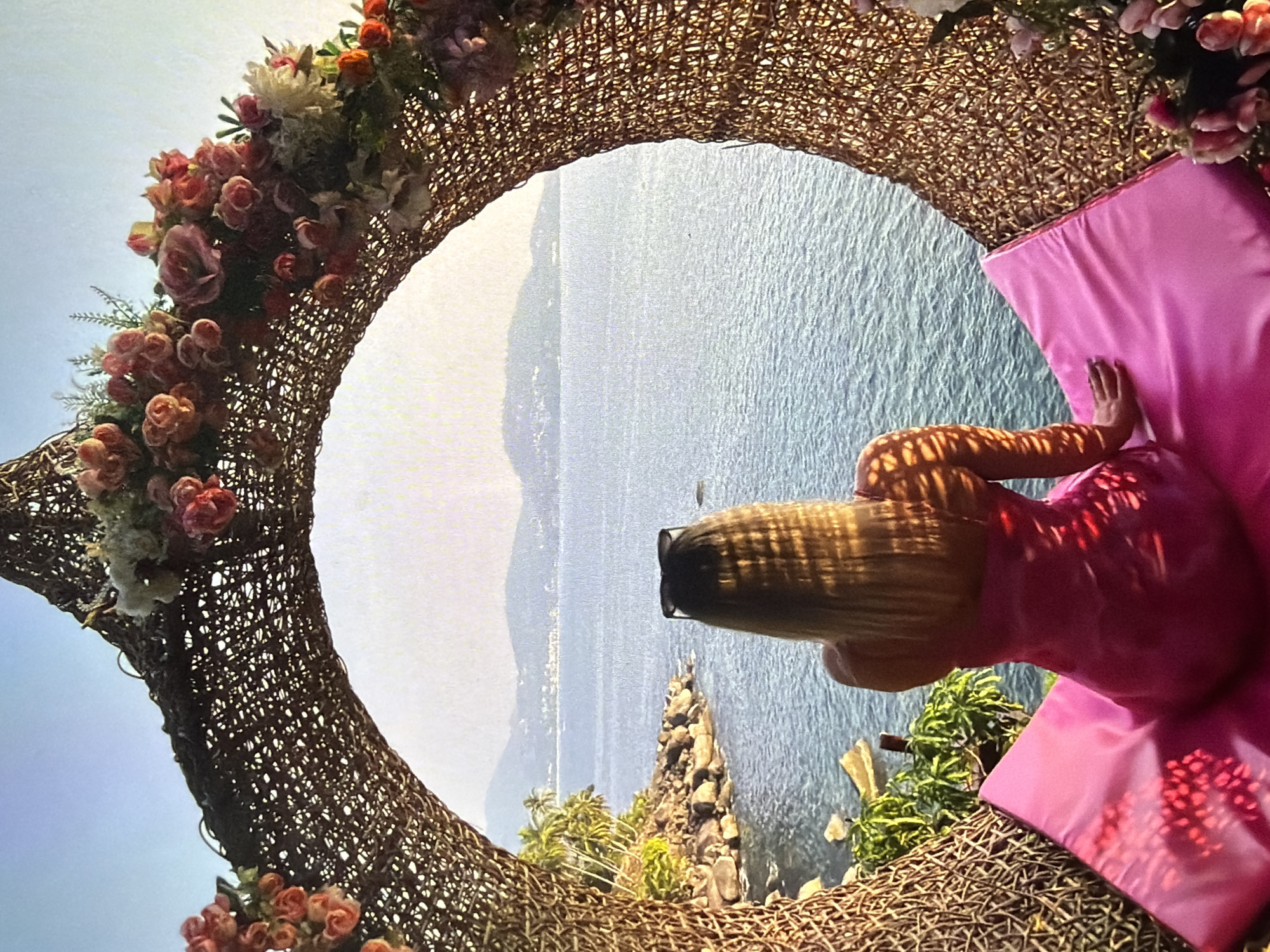 The image captures a person reclining within a large, ornate circular frame overlooking a vast body of water. The subject, seen from the back, has long blonde hair and is wearing a bright pink dress, with sunglasses resting on the back of their head. They are seated comfortably against a soft pink cushion, facing away from the viewer and towards the scenic vista.
The intricate circular frame is woven from a natural material like wicker or rattan and is profusely decorated with artificial flowers and foliage in shades of pink, red, and white. This decorative structure acts as a window, framing the expansive view beyond. The background features a serene body of water, likely an ocean or large lake, with distant mountains or hills faintly visible on the horizon under a bright sky. Below the frame, a rocky coastline with sparse green vegetation, including small trees or shrubs, suggests an elevated or cliffside location.
Bright daylight illuminates the scene, creating noticeable shadows and highlights, particularly visible as dappled patterns on the person's arm and dress, likely cast by the woven frame or nearby foliage. The overall atmosphere conveys relaxation and scenic beauty. The time of day appears to be daytime, with clear weather. No visible text is present in the image.