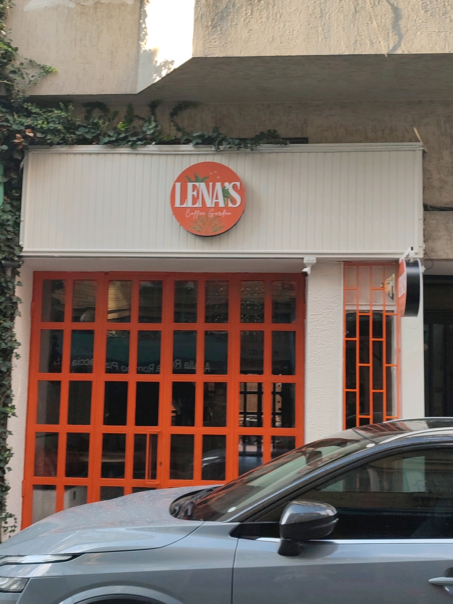 A grey car is parked in front of a building with orange doors. The building has a sign that says "LENA'S Coffee Garden". There is greenery growing on the left side of the building. The scene is located in Kadıköy, Turkey.