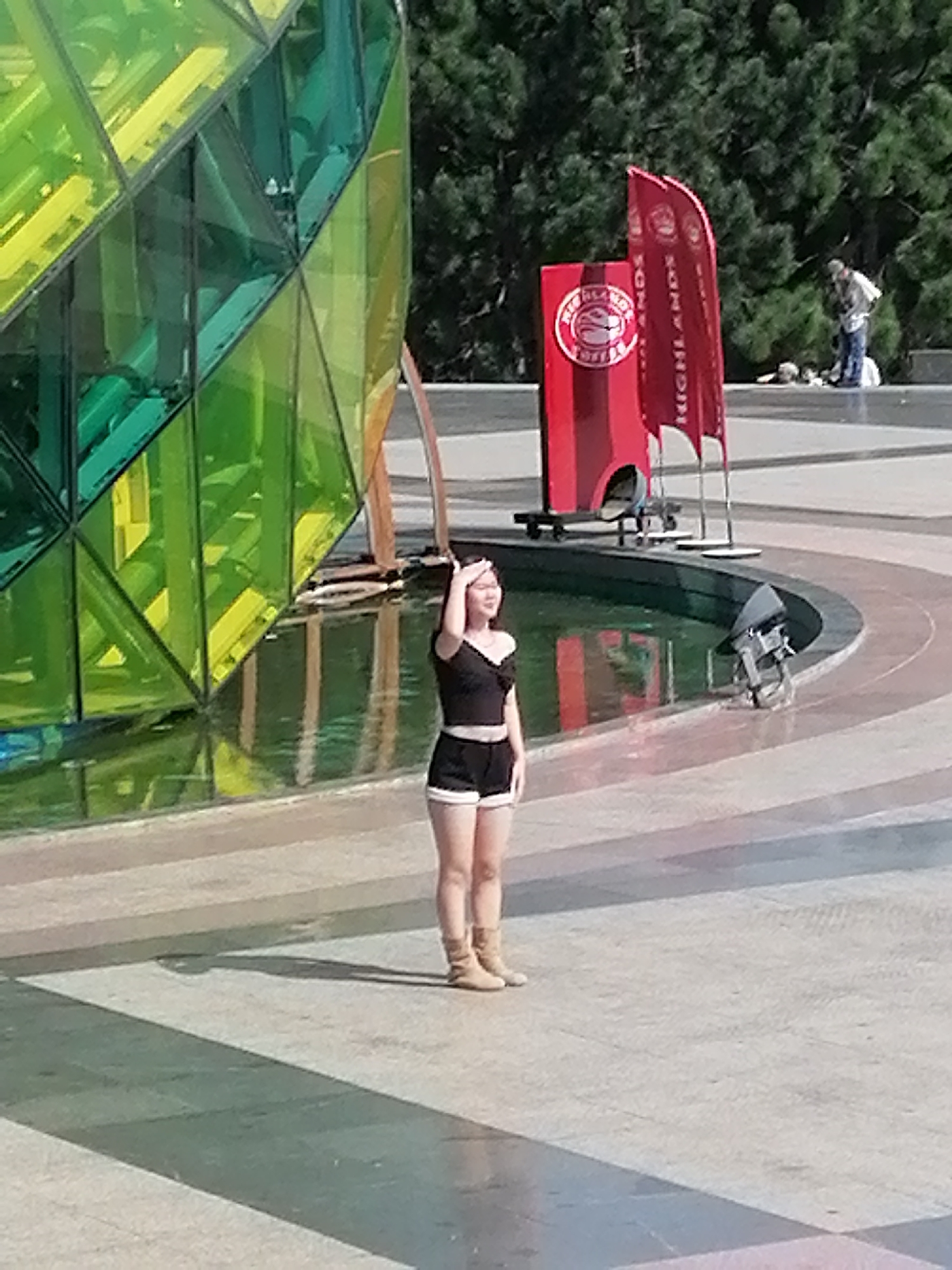 The image depicts an outdoor scene in Da Lat, Vietnam, near a large, faceted glass structure, likely a building or art installation, with green and yellow colored panels. A young woman in a black off-the-shoulder top and black shorts with white trim stands in the foreground, her right hand raised to her forehead as if shielding her eyes from the sun. She is wearing light brown ankle boots. Behind her, to the left, is a circular pool of water reflecting the sky and surrounding structures. To the right of the woman, a red display booth with "HIGHLANDS COFFEE" branding is visible, along with two red feather flags. Further in the background, on a raised walkway, a person in a light-colored shirt and blue jeans is bent over, possibly attending to something on the ground. Pine trees form a dense backdrop. The ground is paved with stone tiles in various shades of grey and tan, arranged in geometric patterns. The lighting suggests it is a sunny day.