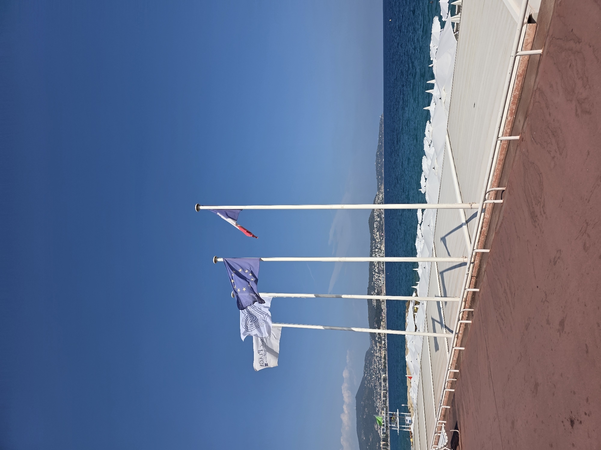 This is a medium-shot, eye-level, vertical, outdoor photograph taken on a sunny day in Nice, France, along the Promenade des Anglais. The foreground features a reddish-brown paved walkway. Behind it, a white metal railing separates the walkway from a raised white platform covered with rows of white umbrellas. Beyond the platform is the blue Mediterranean Sea, and in the distance, a coastline dotted with buildings and green hills under a clear blue sky.
Prominently featured in the middle ground are three tall, white flagpoles. The leftmost flagpole displays a white flag with a logo in the center that appears to be a heart shape formed by two intertwined figures. The text on the flag is partially obscured and reversed, reading "PLAZA." The middle flagpole flies the European Union flag, a blue field with twelve gold stars arranged in a circle. The rightmost flagpole holds the French flag, a tricolor of blue, white, and red vertical stripes, with the red stripe on the right. A fourth, shorter flagpole, partially visible on the far right, appears to fly a small white flag.
A lifeguard tower with a green flag is visible on the left side of the beach area. In the far distance, a lighthouse can be spotted on the peninsula. The overall scene depicts a typical view of the Nice coastline, characterized by its blue waters, sunny weather, and the iconic promenade.