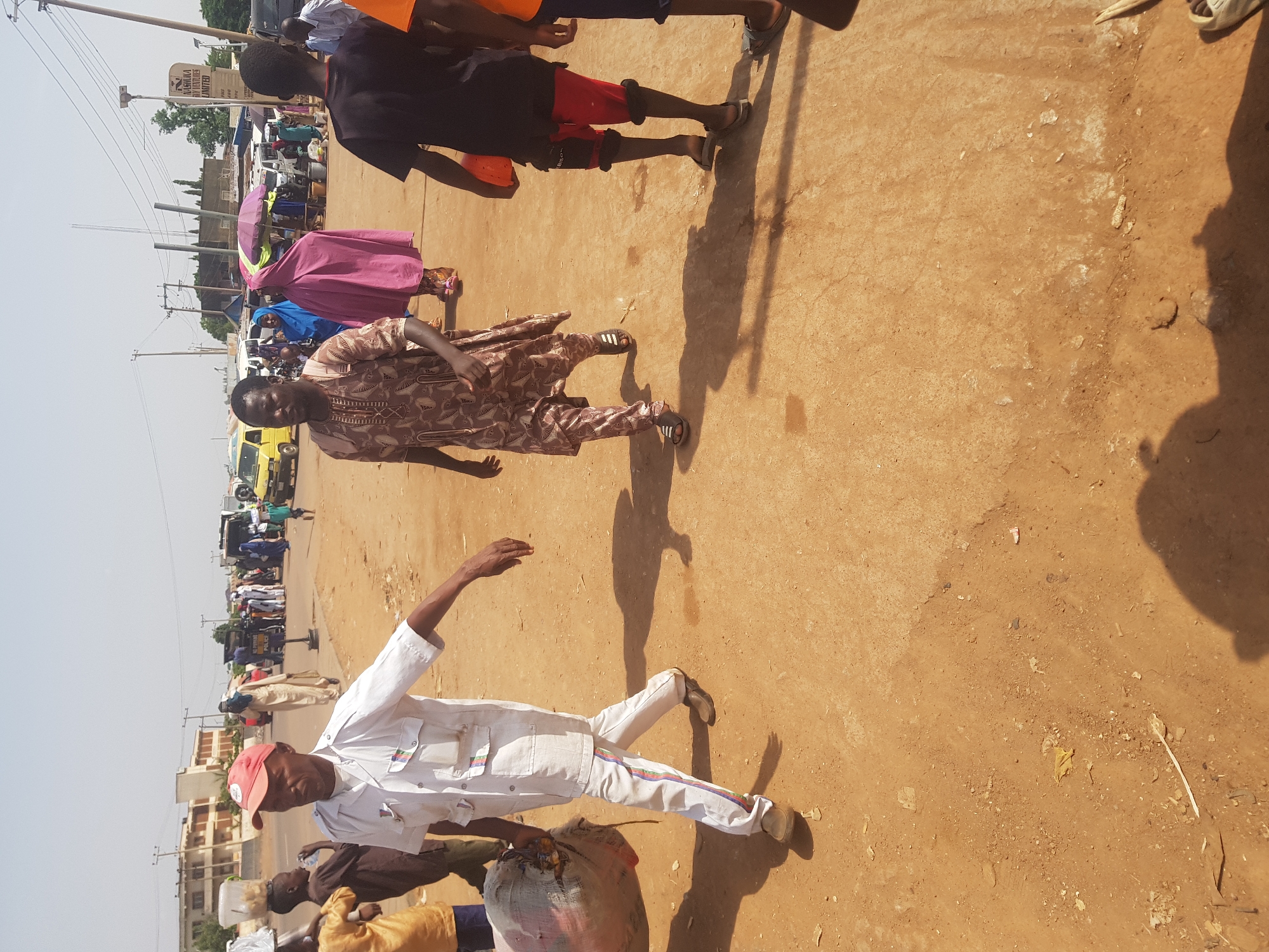 This image captures a bustling outdoor scene on an unpaved street or market area in Tukur Tukur, Nigeria, under bright sunlight. The ground is dry, light brown dirt or sand, showing signs of wear and minor debris. Strong, elongated shadows are cast across the ground, indicating a clear, sunny day, likely during late morning or early afternoon.
In the foreground, a man wearing a white uniform with colorful stripes on the trousers and a red baseball cap is walking towards the left of the frame. He is carrying a large, bulging sack or bag on his left shoulder. Close to him, the head and shoulder of a child, as well as another person's leg and foot, are partially visible.
Further into the midground, another man walks in a brown patterned traditional tunic or robe and trousers, wearing open-toed sandals. Behind him, two figures are visible; one in a vibrant pink dress or robe, holding a purple and green umbrella for shade, and another in a blue garment.
The background shows a busy streetscape with numerous people, several vehicles including a yellow van, and various stalls or small businesses. Utility poles with overhead wires line the street. Buildings with light-colored facades are present, and on one, the text "NASARUKA ANY VENTURES LIMITED" is visible, suggesting a commercial establishment. The scene conveys a sense of everyday activity and commerce in a lively community.