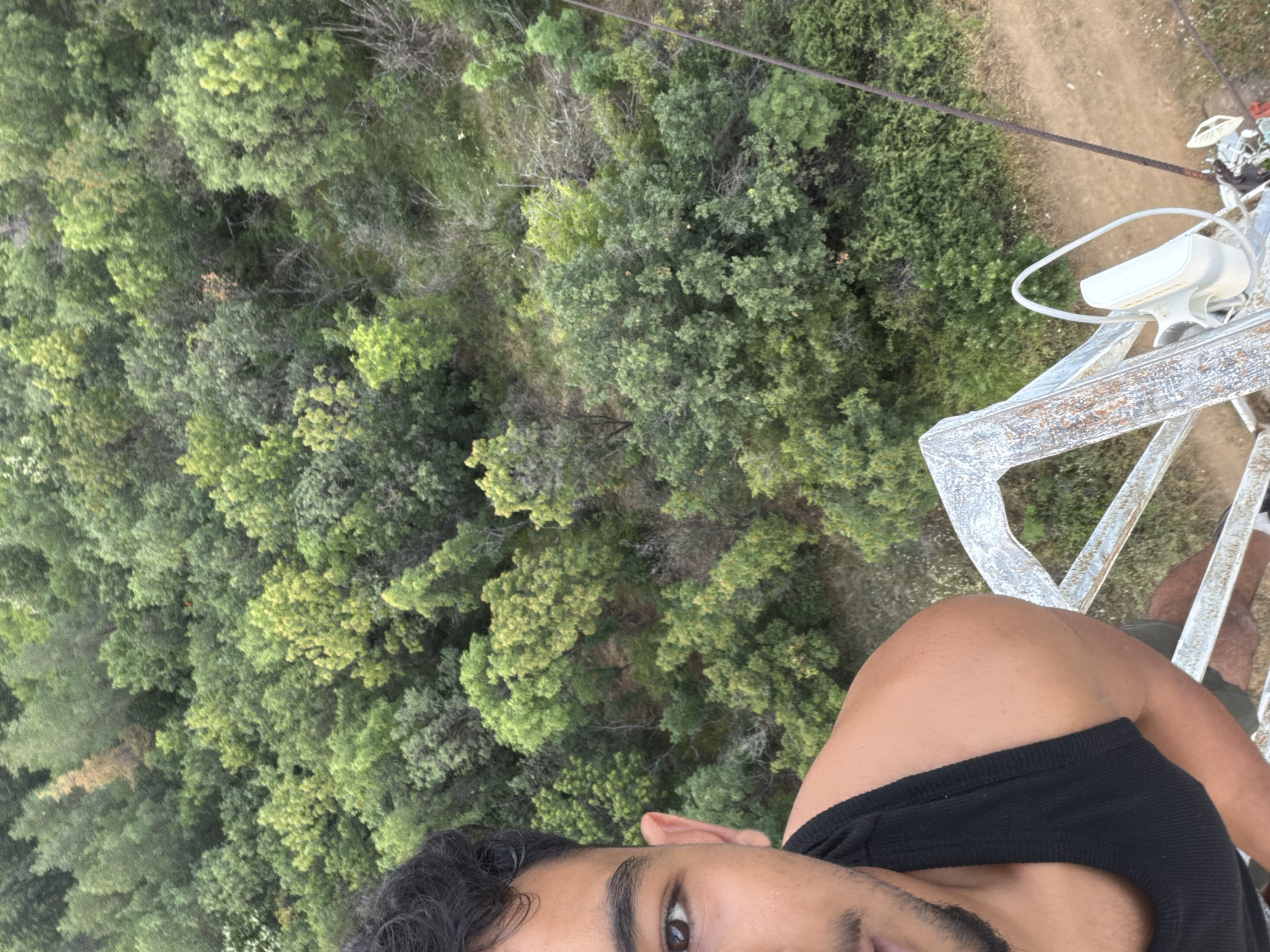 A person is captured in a selfie taken from a high vantage point, looking down at a dense forest and a dirt road. The person is wearing a black tank top and is partially visible in the foreground, with their shoulder, arm, and a portion of their face in view. They have dark hair and a warm skin tone. The background is dominated by a lush green canopy of trees, suggesting a natural, possibly mountainous or hilly, environment. A metallic structure, likely part of a tower or observation deck, is visible to the right, with a white object attached to it that resembles a light fixture or sensor. A thick cable runs diagonally across the upper part of the image, indicating the presence of some form of infrastructure, perhaps related to the tower. The lighting suggests it's daytime, and the overall scene evokes a sense of adventure or exploration, perhaps from an outdoor activity like zip-lining or visiting an elevated viewpoint.