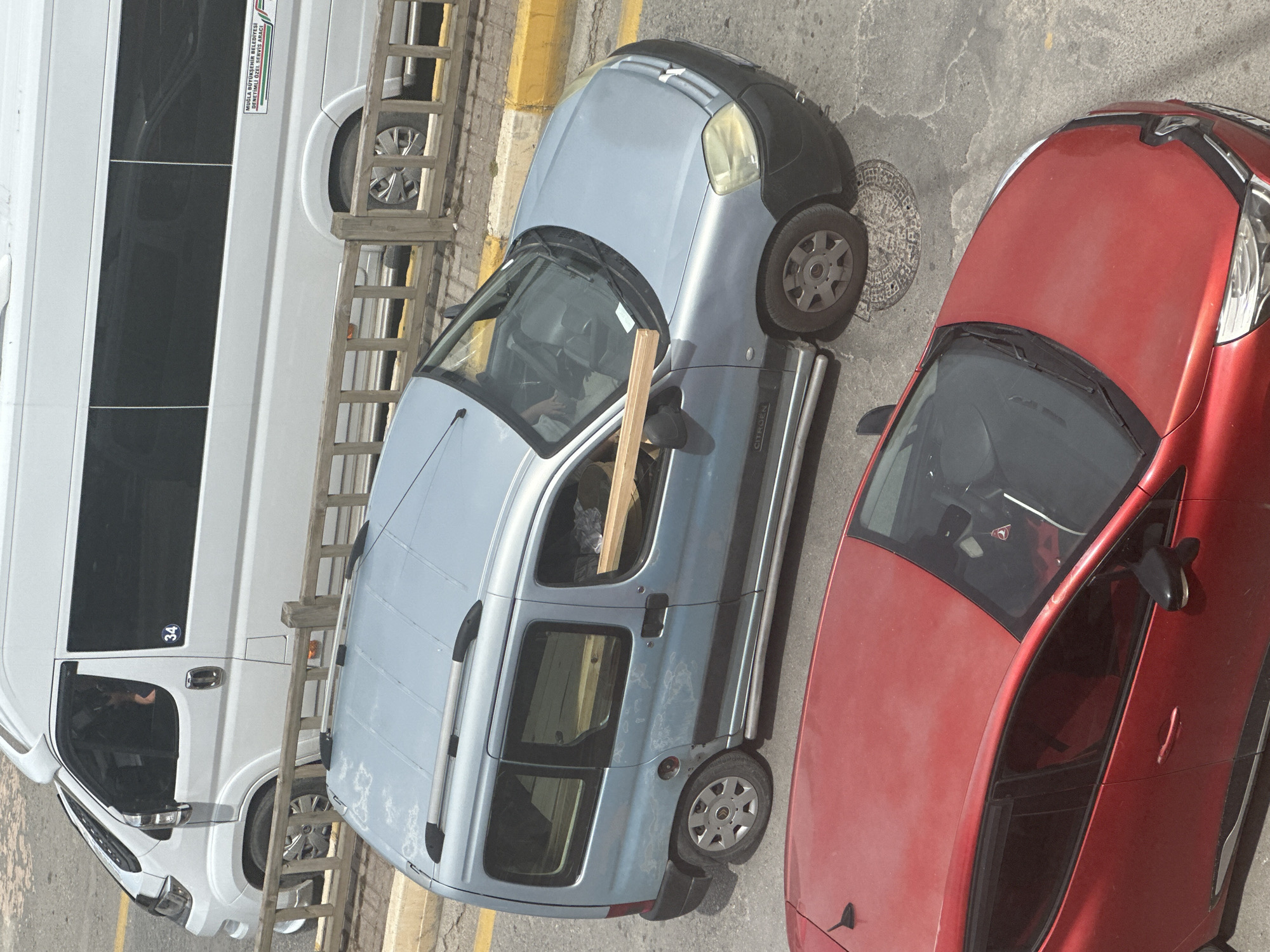 The image shows an overhead view of a street in Fethiye, Turkey. A white van is parked on the left, with the number "34" visible. Next to the van is a blue Citroen Berlingo, with a piece of wood visible inside. A red car is parked to the right of the Citroen. The road surface is concrete, and there is a yellow curb visible. There is a manhole cover behind the Citroen.