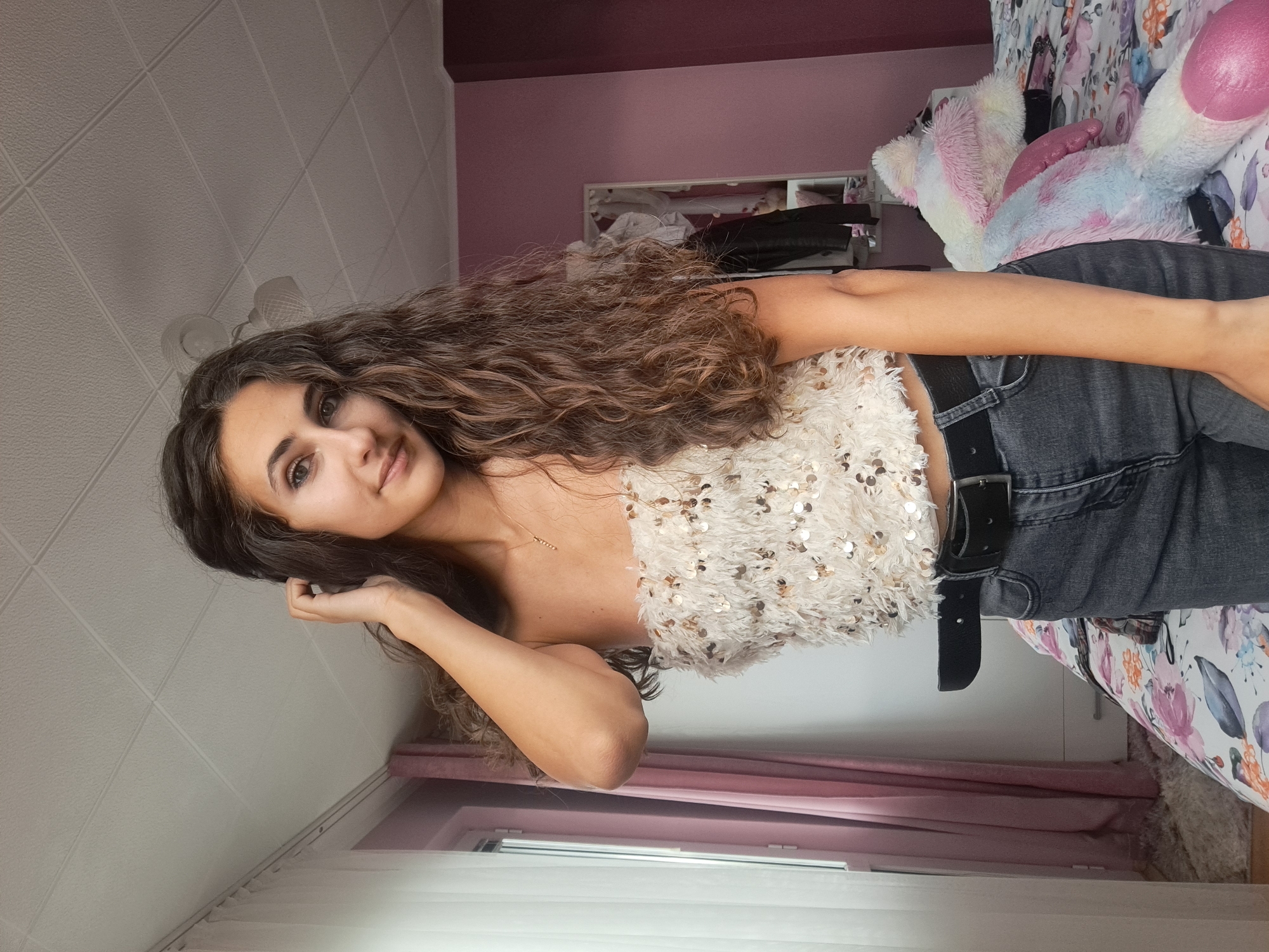 The image shows a young woman with long, curly brown hair standing in a bedroom. She is wearing a sparkly, off-the-shoulder top and dark jeans with a black belt. Her left hand is resting on her head. A large stuffed animal, predominantly pink, blue, and white, is visible on the bed behind her. The room has pink walls and a white ceiling with visible paneling. A window with pink curtains is to the left of the woman. There is a mirror visible in the background, reflecting a closet with clothes. The image is a selfie taken from chest level up. There is no visible indication of the location being in the city of Negovan, Bulgaria.