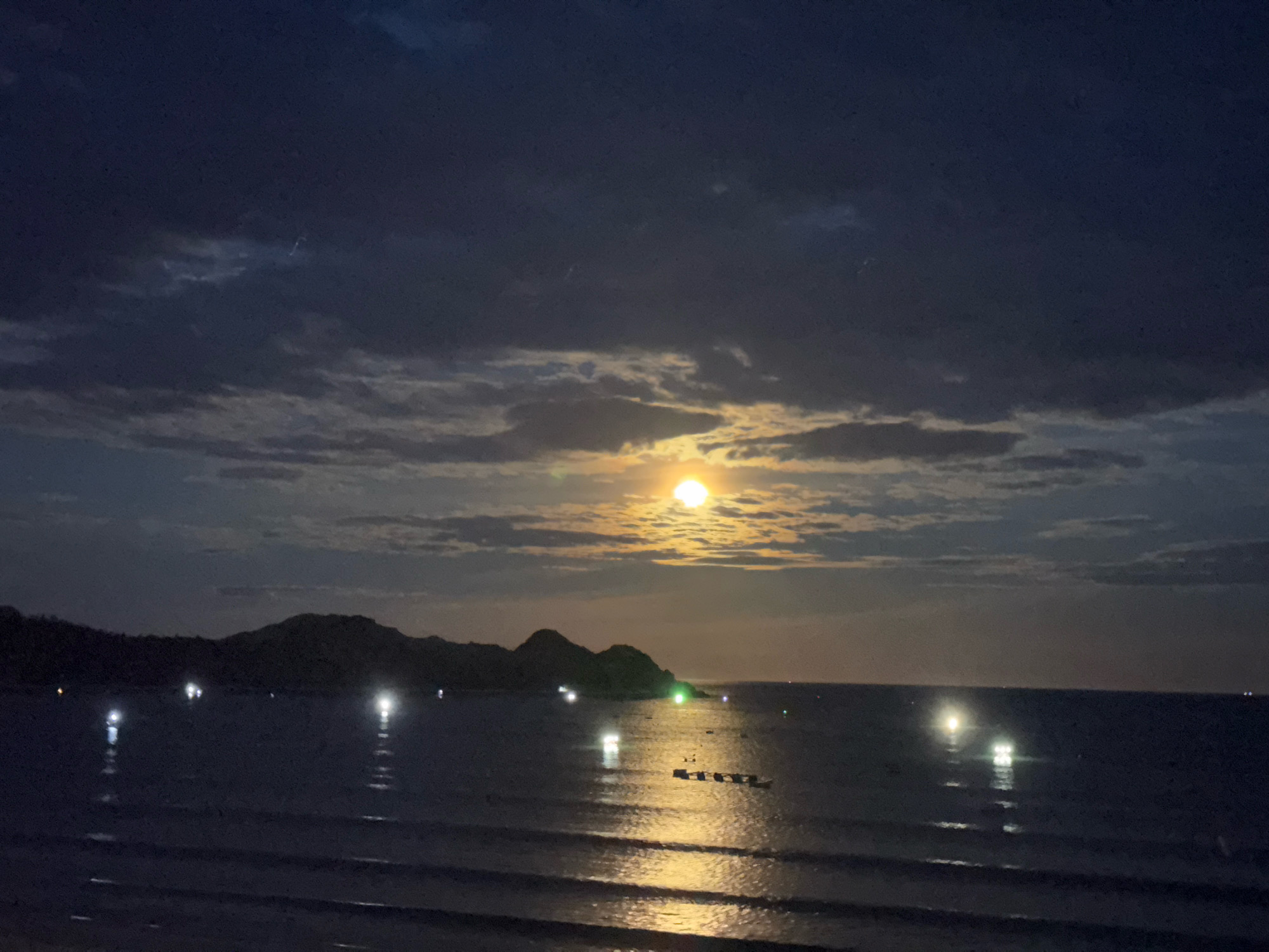 A nighttime photograph captures a serene seascape under a prominent, bright full moon. The moon, a luminous orb, is partially obscured by wispy, dark clouds, casting a warm, golden glow on the surrounding sky. The moonlight creates a shimmering path across the dark water, reflecting the lights of distant boats and the silhouettes of a hilly coastline. Scattered lights, appearing as small, bright points, are visible on the water's surface, suggesting the presence of anchored vessels or buoys. The coastline is characterized by undulating hills, shrouded in darkness, with a few faint lights dotting the landscape. The foreground shows gentle waves lapping at the shore, their surfaces reflecting the ambient light and contributing to the tranquil atmosphere. The overall scene evokes a sense of peace and natural beauty.