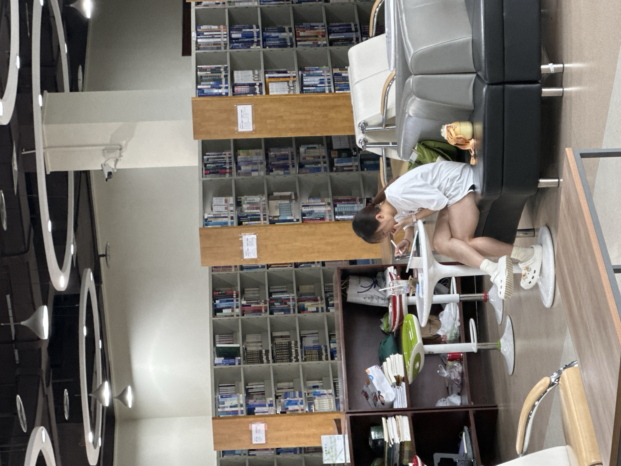 The image shows the interior of what appears to be a library, based on the multiple shelves filled with books. A young woman, wearing a white shirt and dark shorts, is seated on a white chair and is likely studying or reading. She is wearing white socks and Crocs-style shoes. A black sofa is located next to her. There is a brown cabinet adjacent to the chair with various items in it, some of which look like books and other personal belongings.
The location is probably in the city of Chongqing High-tech Zone, China.