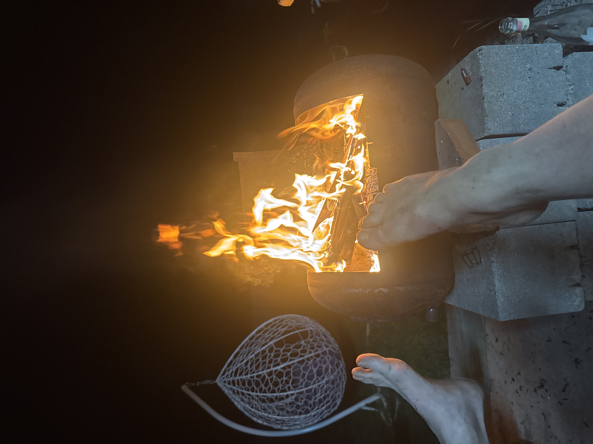 A nighttime scene shows a cylindrical metal fire pit with flames visible inside. A hand is reaching towards the fire pit, while bare feet are visible in the lower part of the frame. A decorative mesh ball hangs from a curved metal frame. The fire pit is supported by concrete blocks on the right side of the image, on which a glass bottle sits. The location is not discernible as Chaikovsky, Russia from the image.