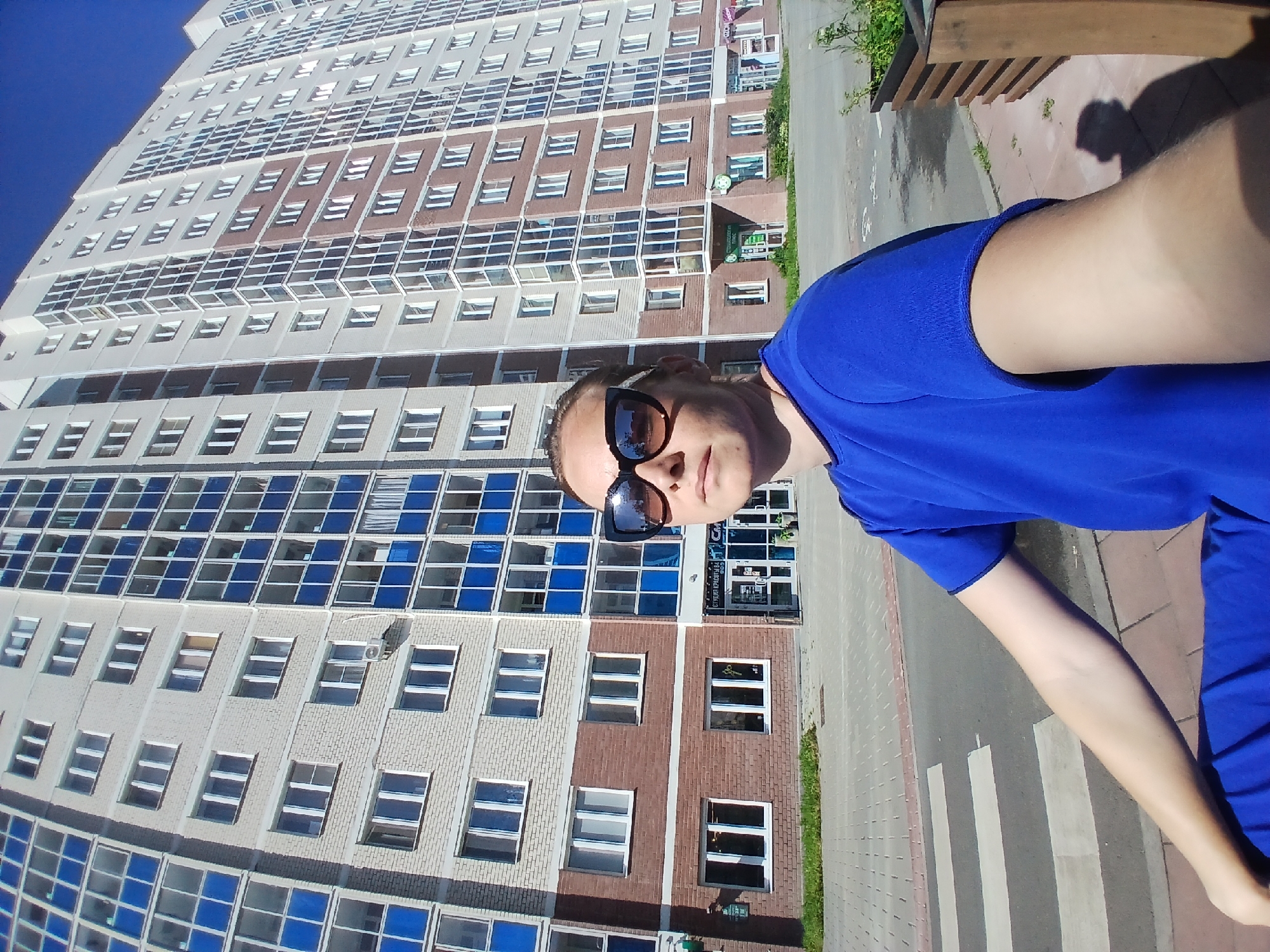 A low-angle, wide outdoor selfie taken on a sunny day shows a woman in a bright blue top and blue pants standing on a pedestrian crossing in front of a modern, multi-story apartment building. She is wearing large, black sunglasses that reflect the building. The building is predominantly light gray brick with numerous windows, some of which are recessed and have blue frames. The lower floors of the building feature red brickwork and storefronts. The sky is clear and blue. The pedestrian crossing is marked with white stripes on gray pavement. The background includes some greenery and a wooden planter box. The image quality is moderately high, with good detail and color saturation. The woman's arm is extended as she takes the selfie, and her body is partially visible in the foreground.