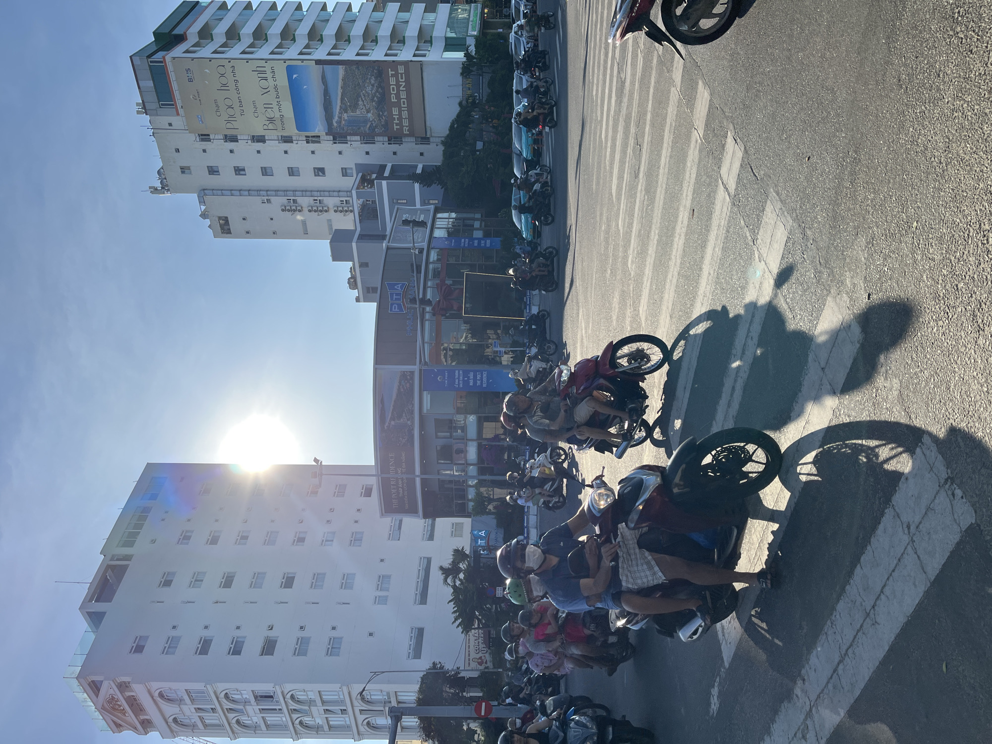 The image depicts a bustling street scene in Đà Nẵng, Vietnam, likely at an intersection. Numerous motorbikes are present, many carrying multiple passengers, indicating heavy traffic. Several tall buildings flank the street, one displaying an advertisement for "The Poet Residence" and "Biển xanh." The buildings suggest an urban environment, possibly a residential or commercial district. Sunlight glares between the buildings. A crosswalk is visible, and a few pedestrians are visible alongside the road.