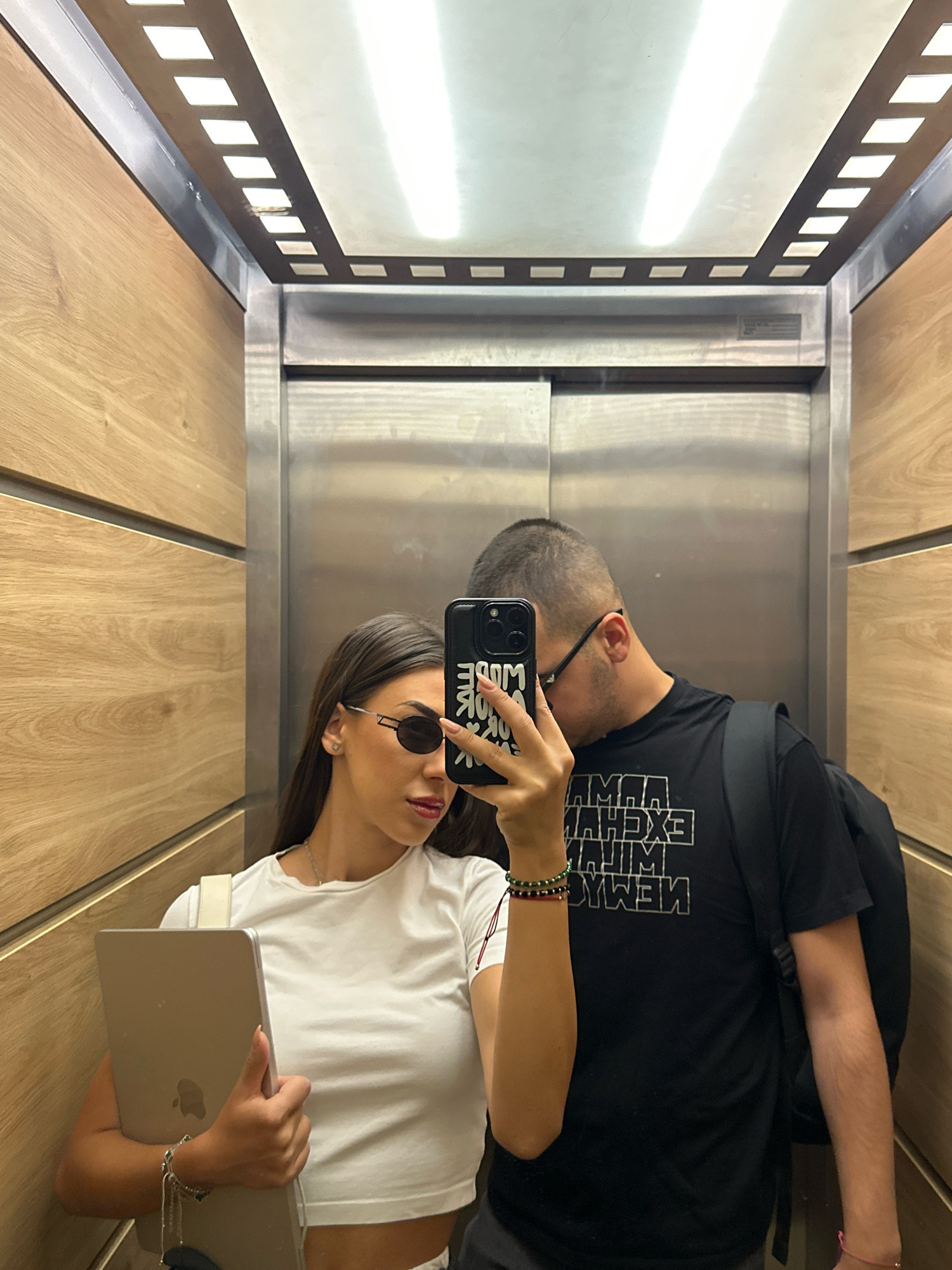 A young couple is taking a selfie in an elevator. The woman, on the left, is wearing a white crop top, sunglasses, and holding a laptop. She has dark hair and is wearing bracelets on her wrist. The man, on the right, is wearing a black t-shirt with white text and a backpack. He has short hair and is wearing glasses. The elevator has wood-paneled walls and a metal door. The lighting in the elevator is provided by fluorescent lights in the ceiling. The scene is not indicative of any specific location in Sofia, Bulgaria.