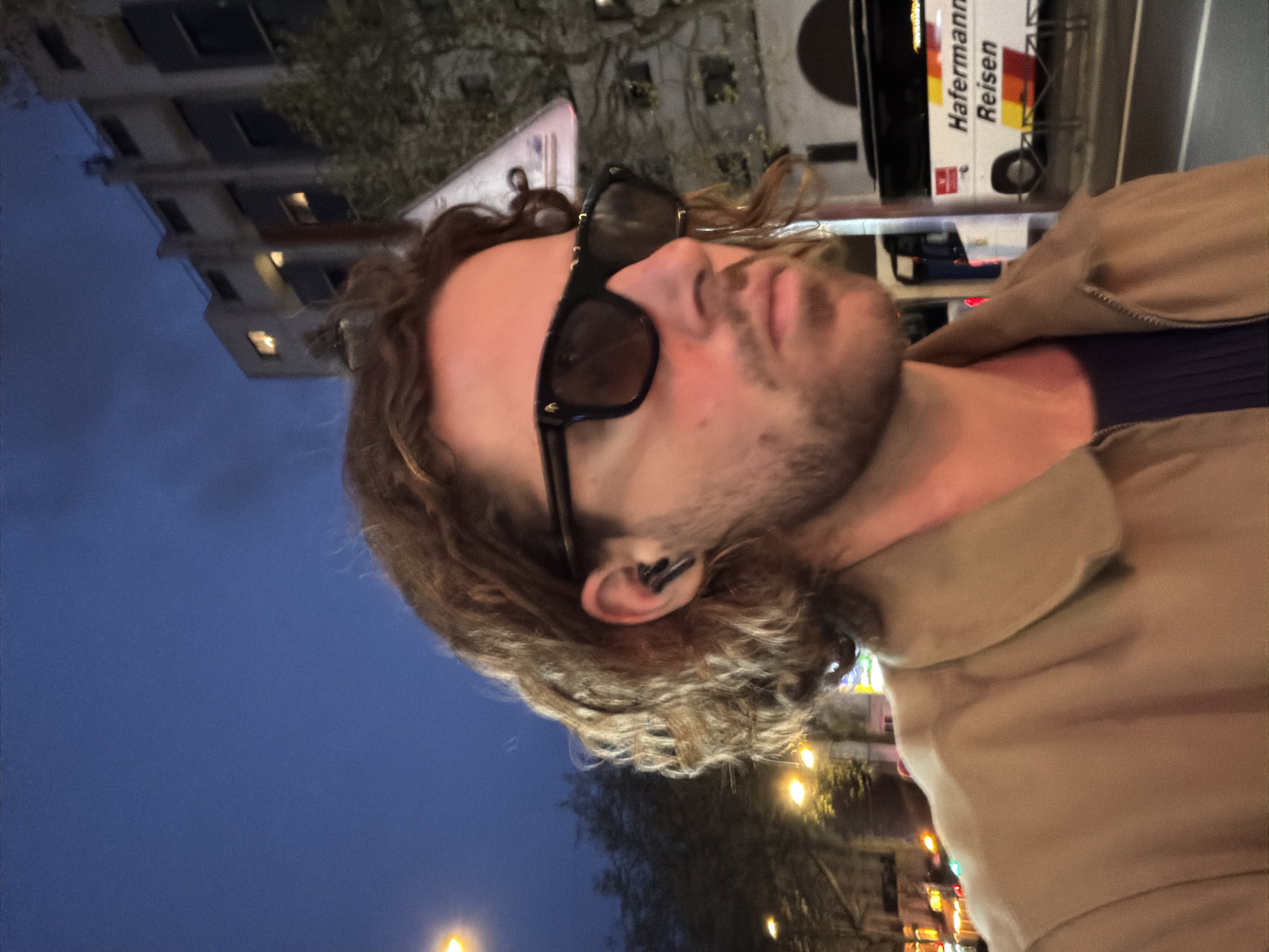 The image is a portrait of a Caucasian male with curly brown hair and a beard, wearing sunglasses and an earbud in his left ear. He is wearing a brown jacket. In the background, a building with illuminated windows suggests a city environment. A bus with the text "Hafermann Reisen" is visible. The presence of trees and lights implies an urban street setting. Based on the architectural style and the text on the bus, the location may be near Paris, France. The time appears to be evening or night, given the dark sky and artificial lighting.
